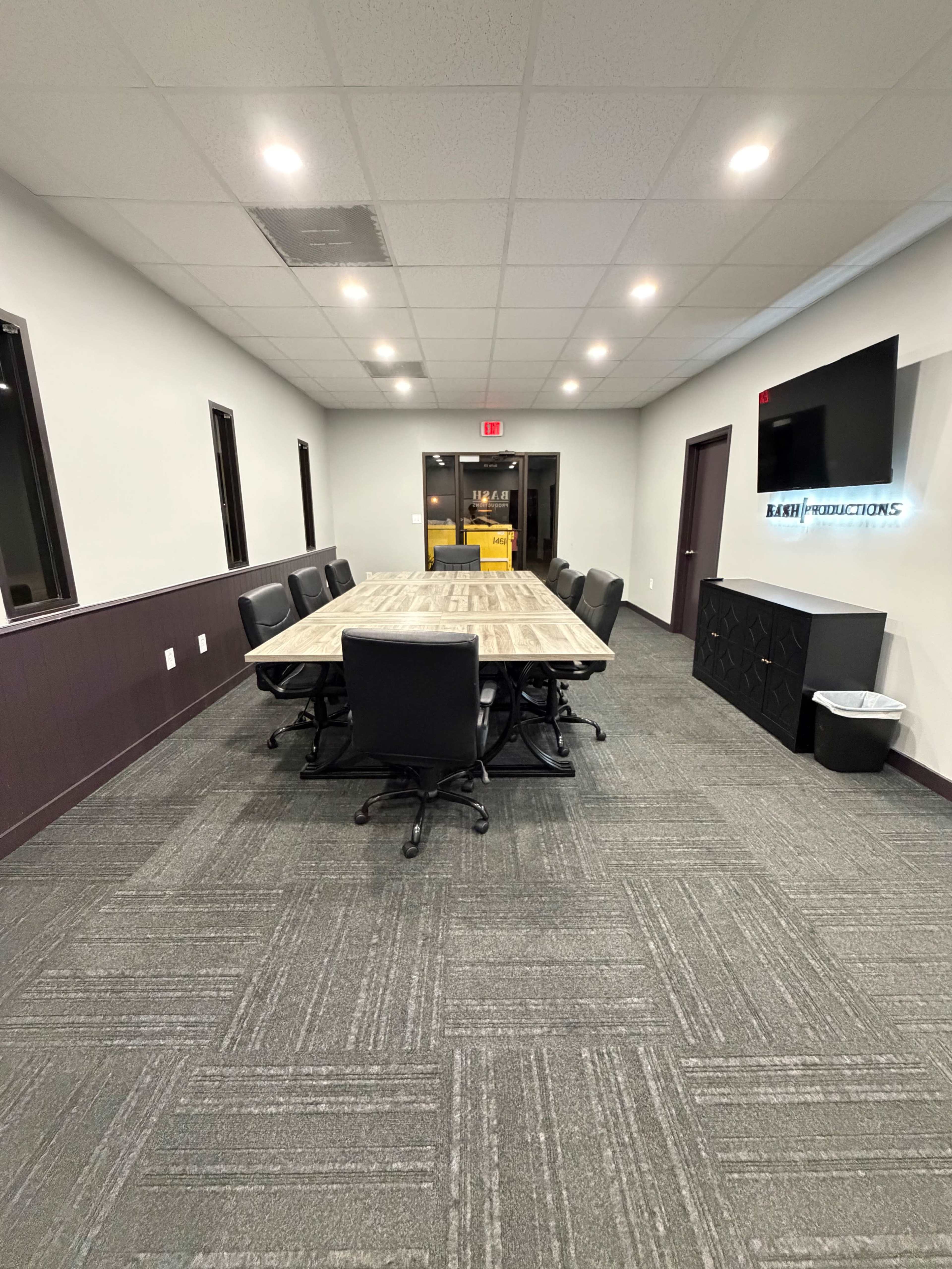 A conference room features a large wooden table surrounded by black chairs, with a wall-mounted screen and sliding glass doors at one end.