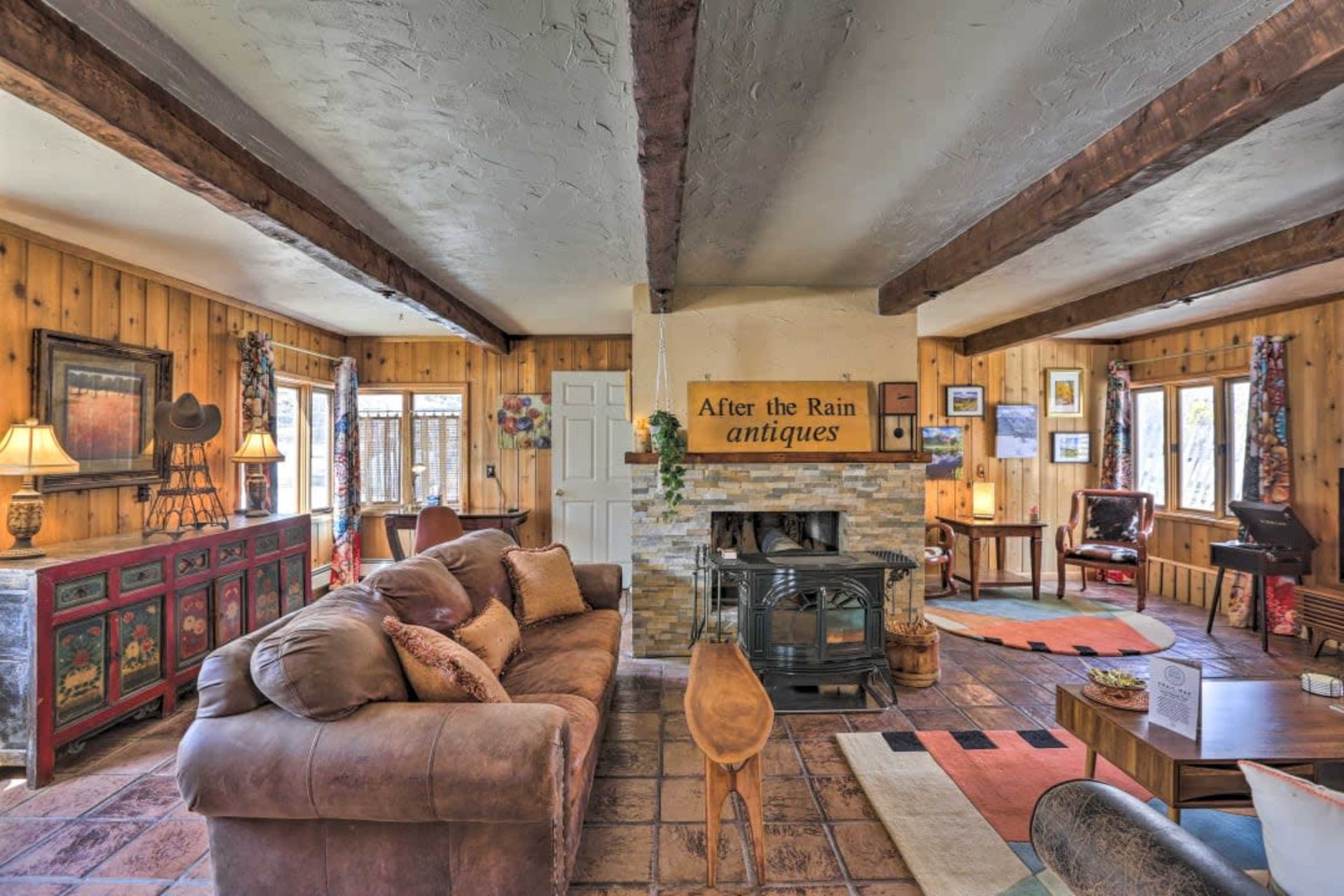 A rustic living room featuring a large brown sofa, a stone fireplace, wooden beams, and antique decor against a background of warm wooden walls.