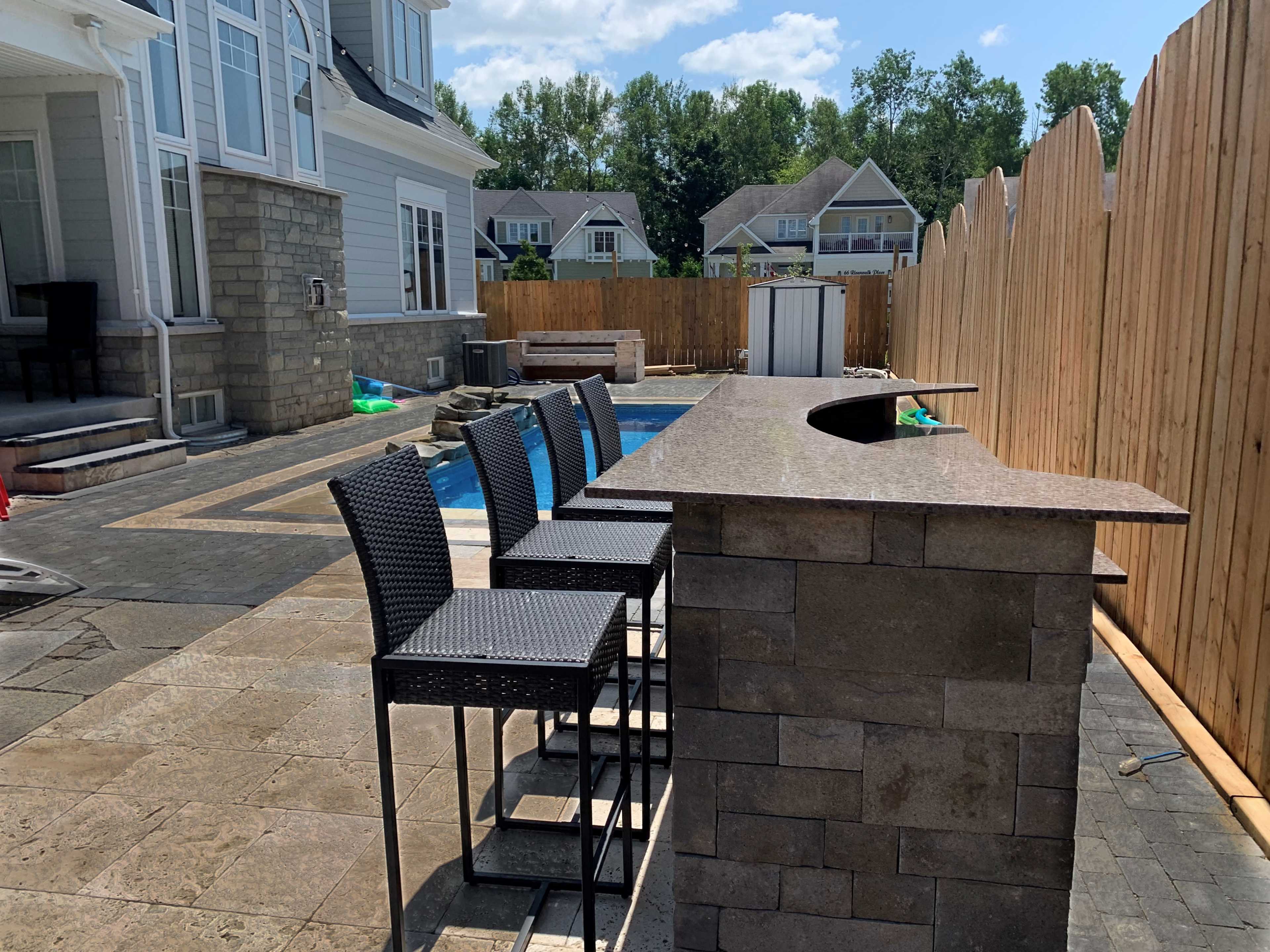 An outdoor patio area featuring a stone bar with high stools, next to a swimming pool surrounded by a wooden fence.