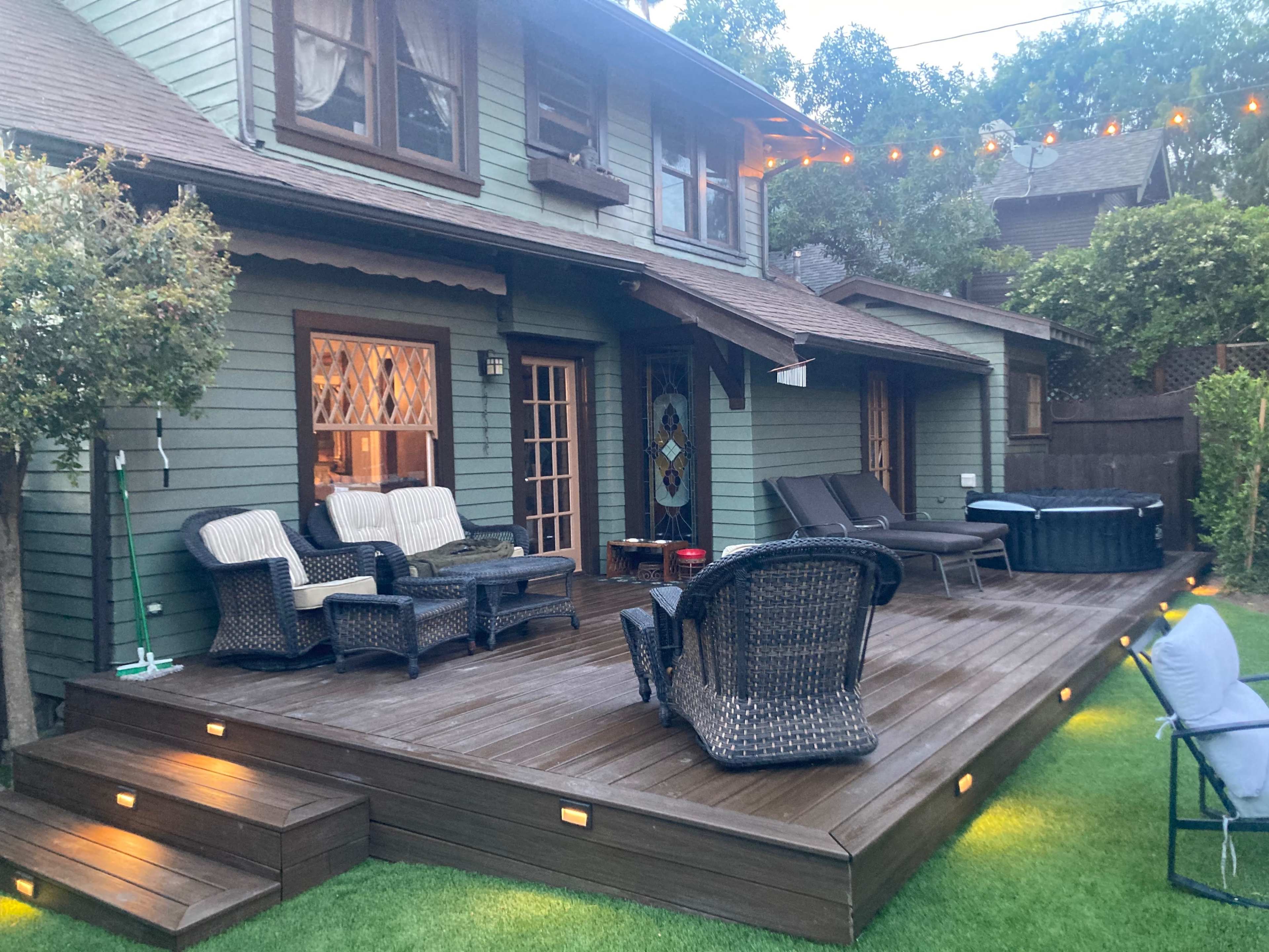 The image shows a two-story house with a wooden deck featuring lounge chairs, outdoor seating, and a hot tub, surrounded by greenery and lighted string decorations.