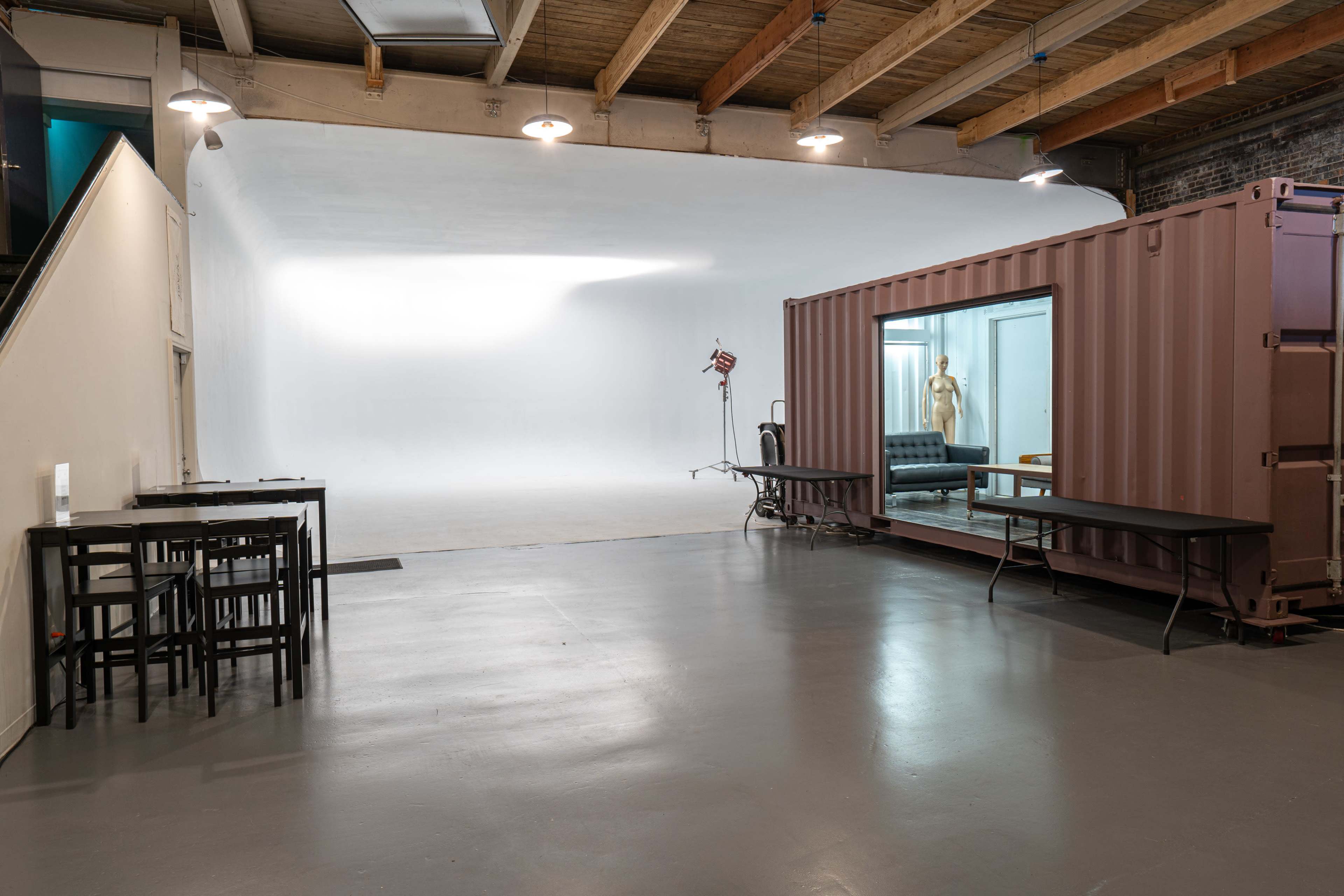 An industrial studio space featuring a white backdrop, a shipping container converted into a room, and a few tables and chairs arranged nearby.