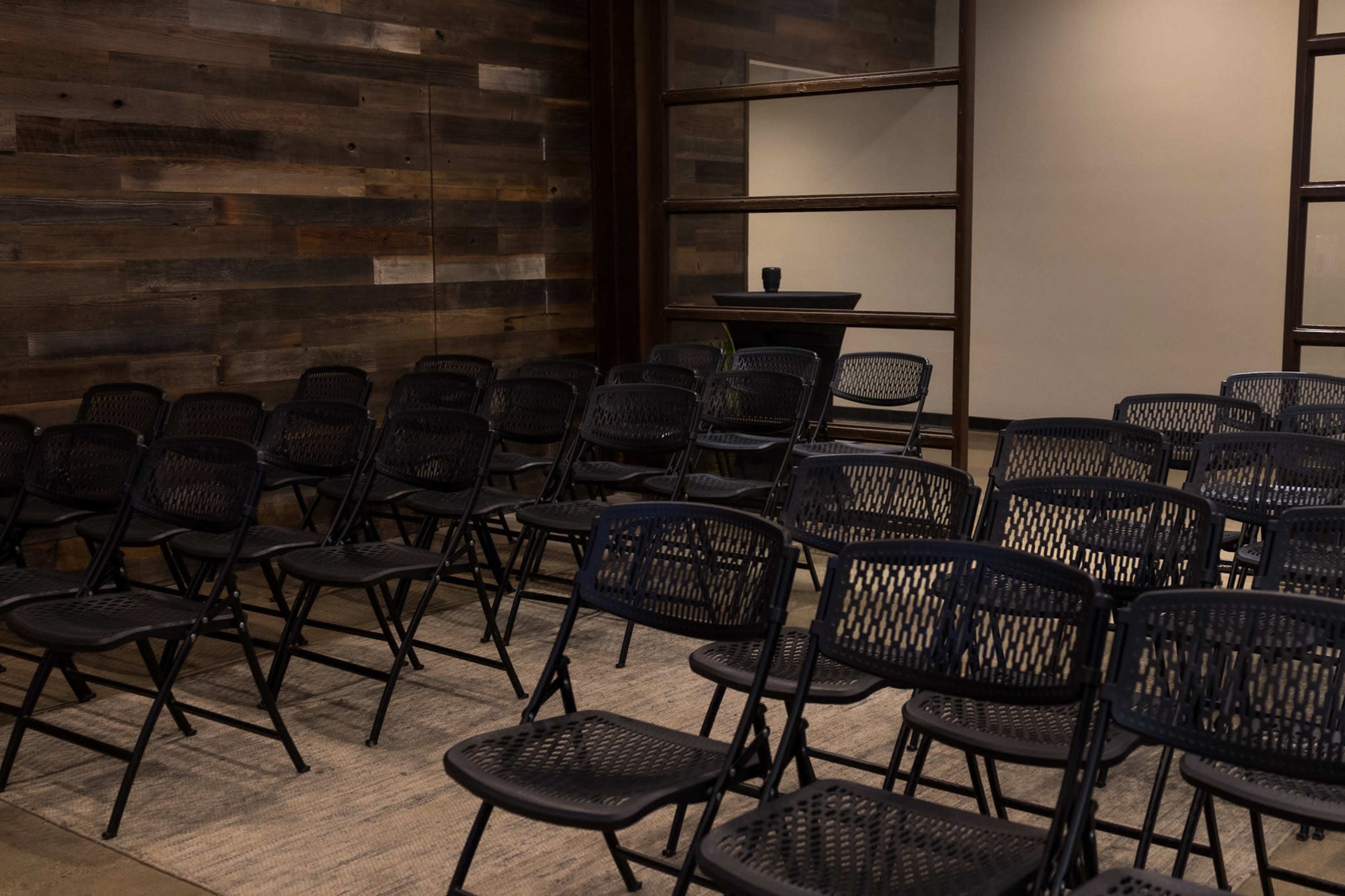 A row of black folding chairs is arranged in a room with wood-paneled walls and a small table at the back.