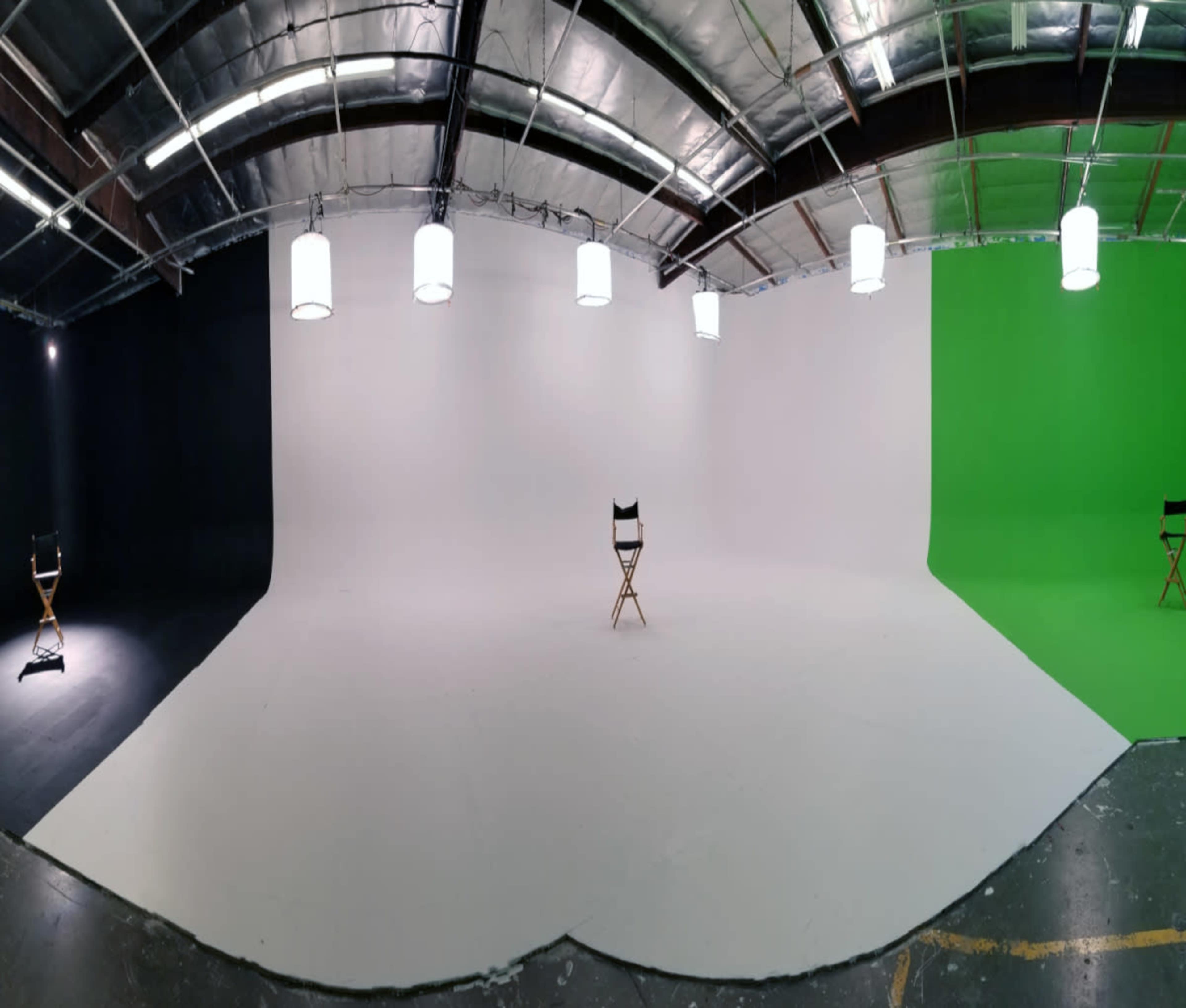 A large, empty studio space with a black wall on one side, a white wall in the center, and a green wall on the opposite side, all featuring a single wooden director's chair positioned in front of the white backdrop.