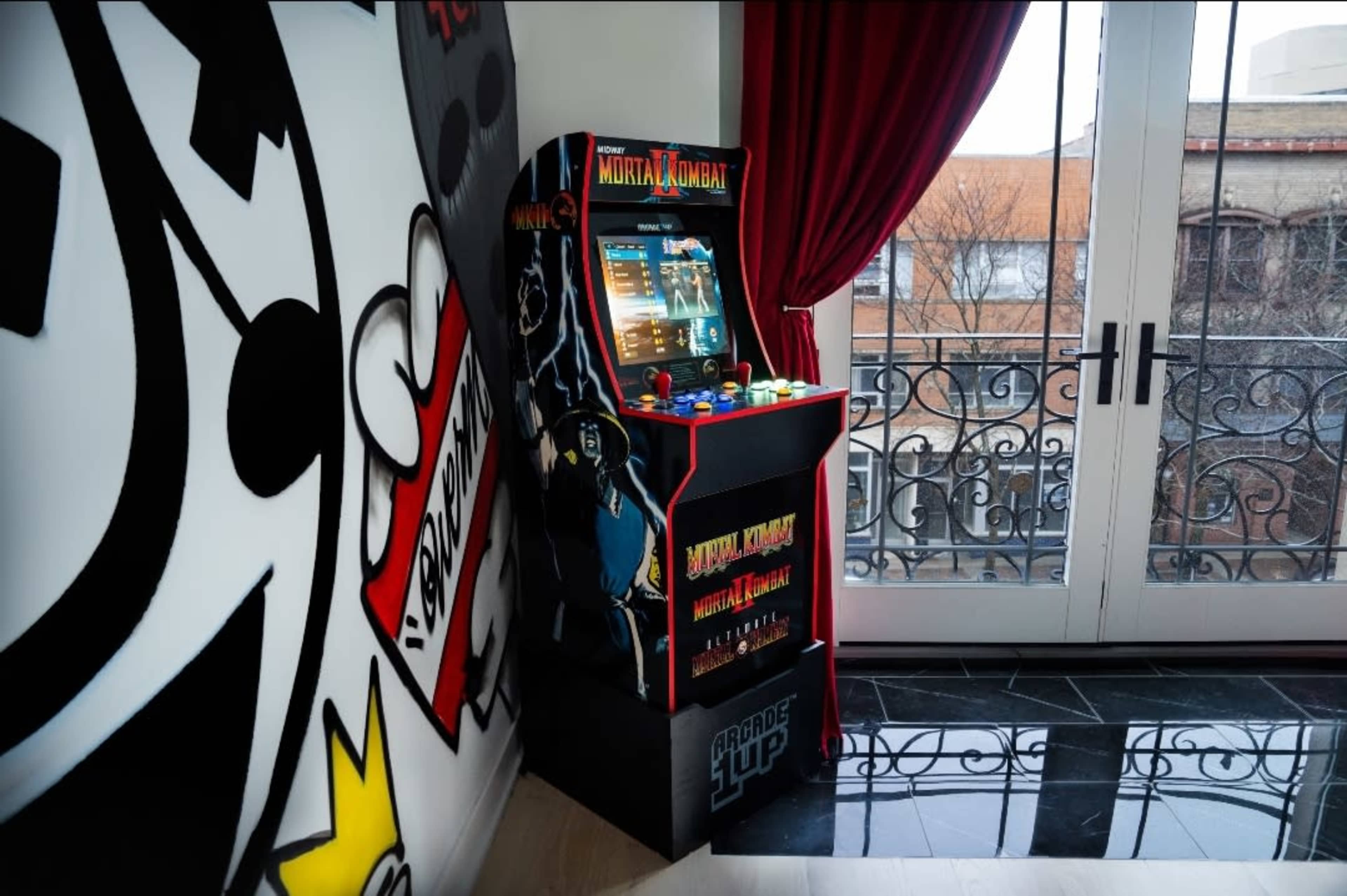 A Mortal Kombat arcade machine stands next to a large window with iron railings and red curtains in a modern room.