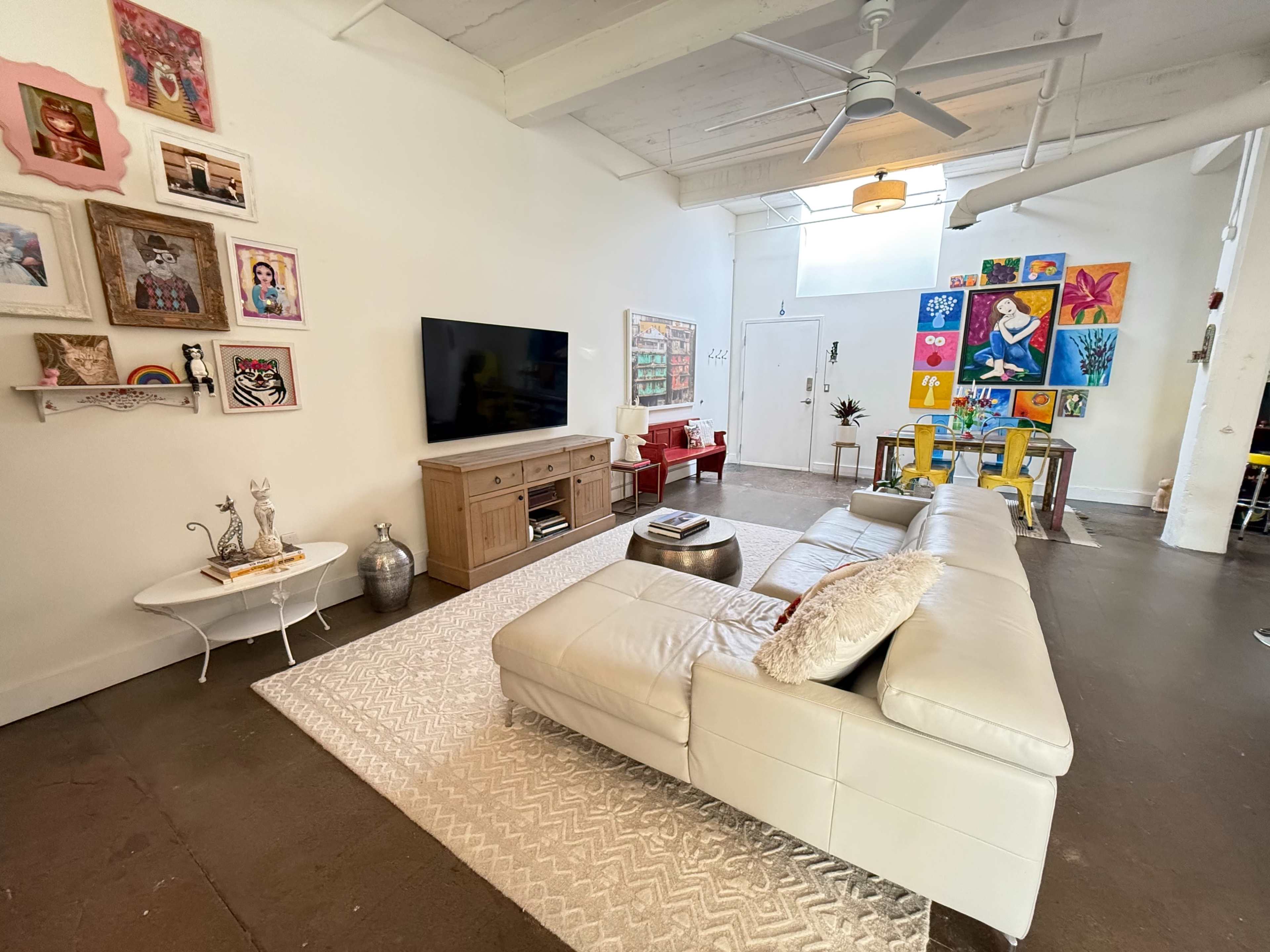 The image shows a modern living room with a sectional white leather sofa, a wooden TV stand, and colorful artwork on the walls.