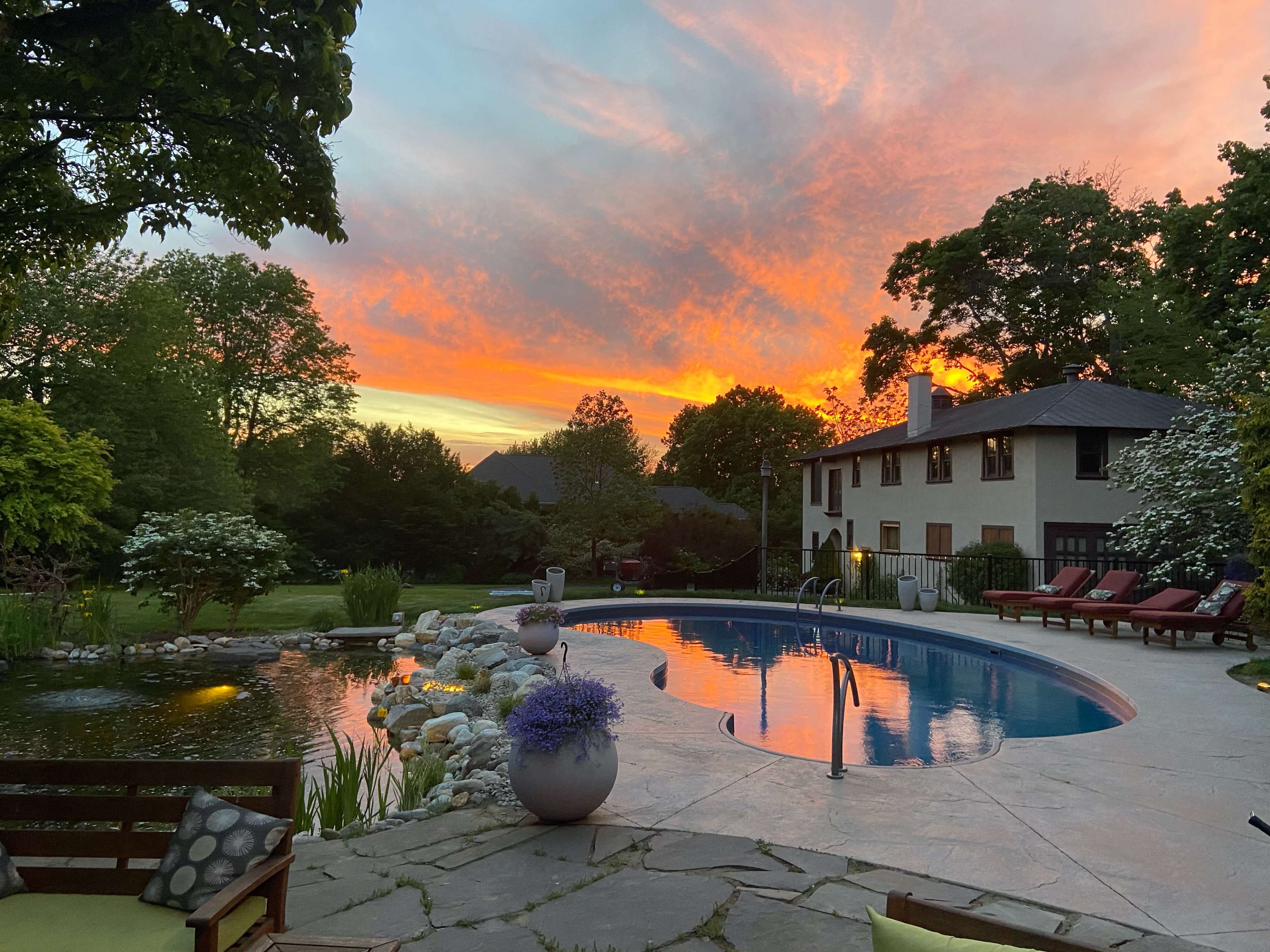 The scene features a swimming pool surrounded by lounge chairs, with a vibrant sunset illuminating the sky in the background.