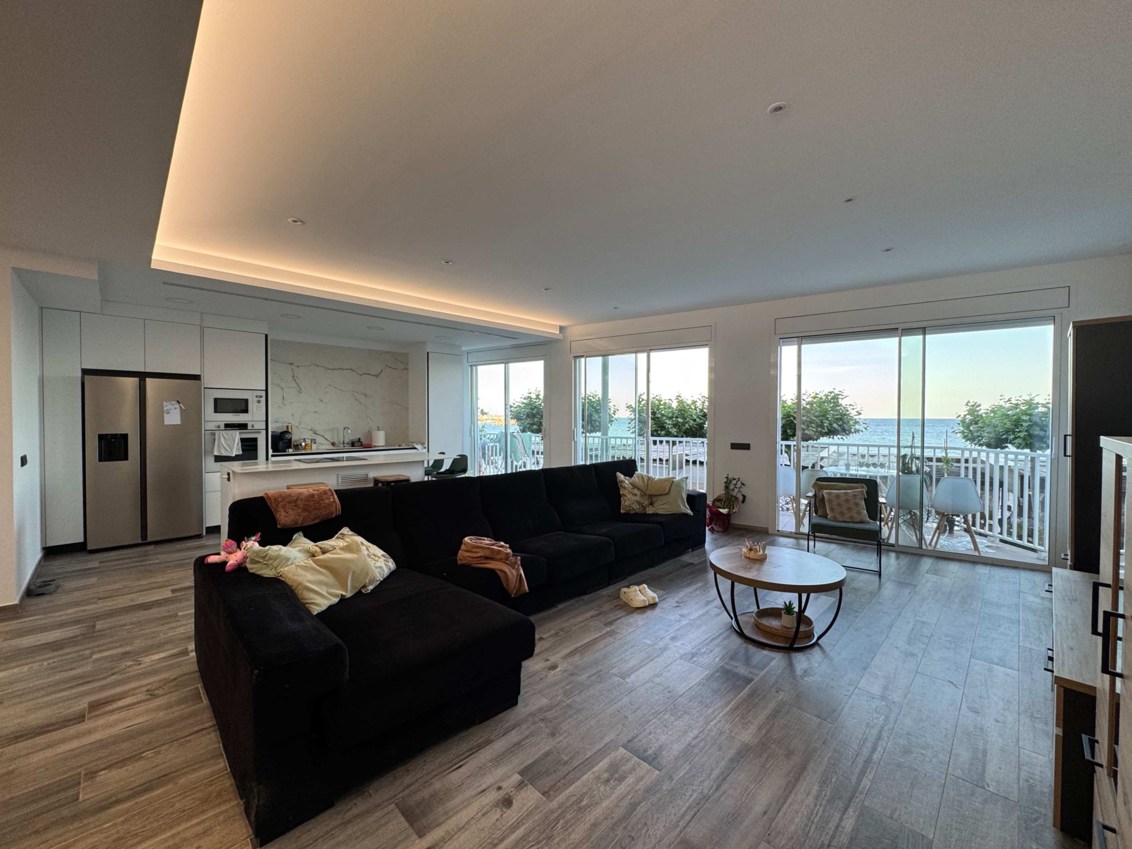 A spacious living room features a black sectional sofa, a round wooden coffee table, and large windows with a view of the outdoors.