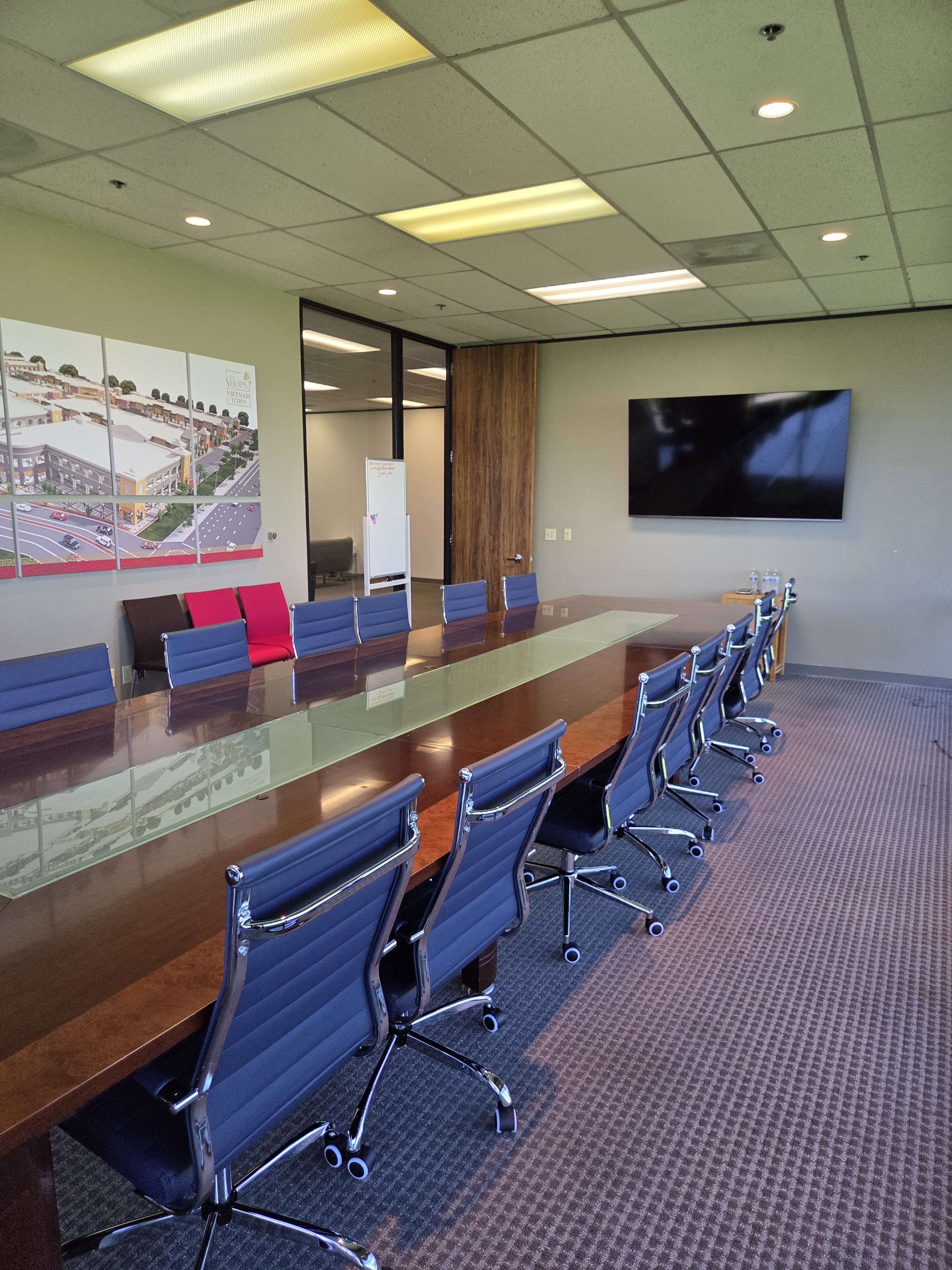 A modern conference room features a long wooden table surrounded by structured black office chairs, with a large screen on one wall and informational displays on another.