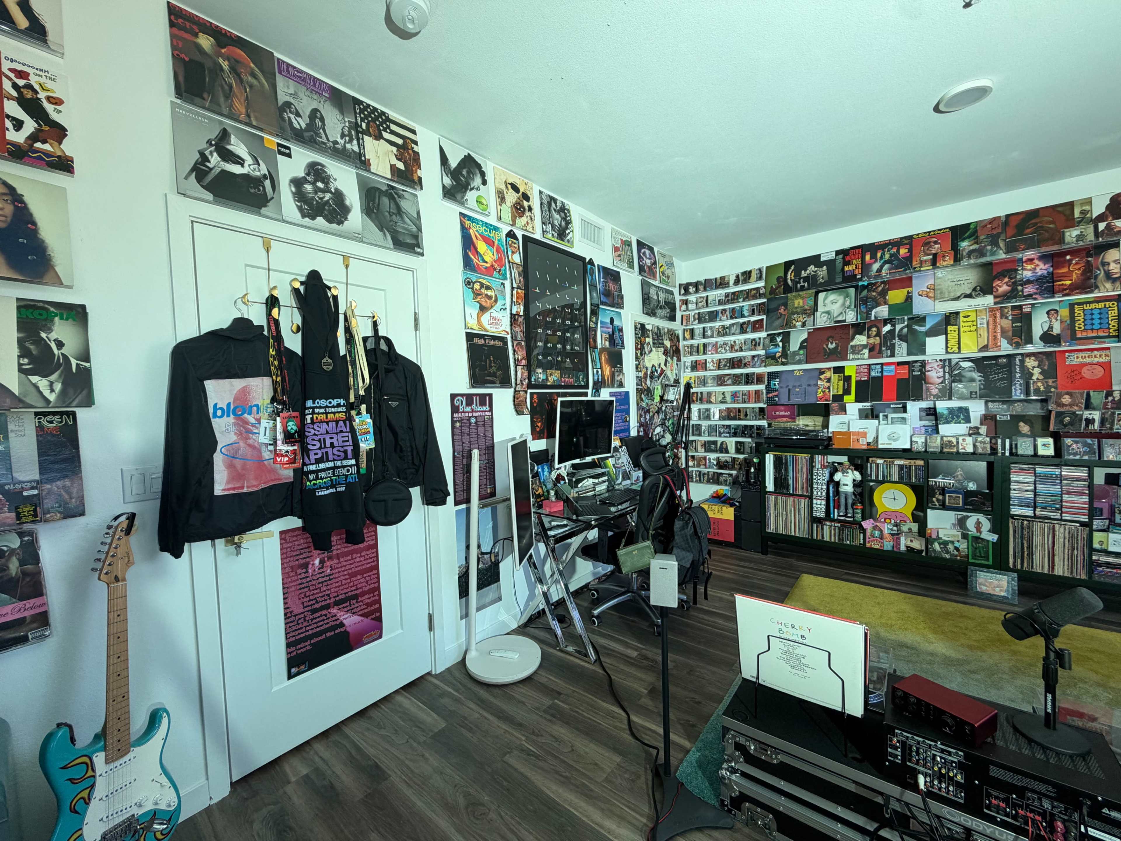 The image shows a room filled with music memorabilia, including posters on the walls, a computer setup, and various musical instruments.