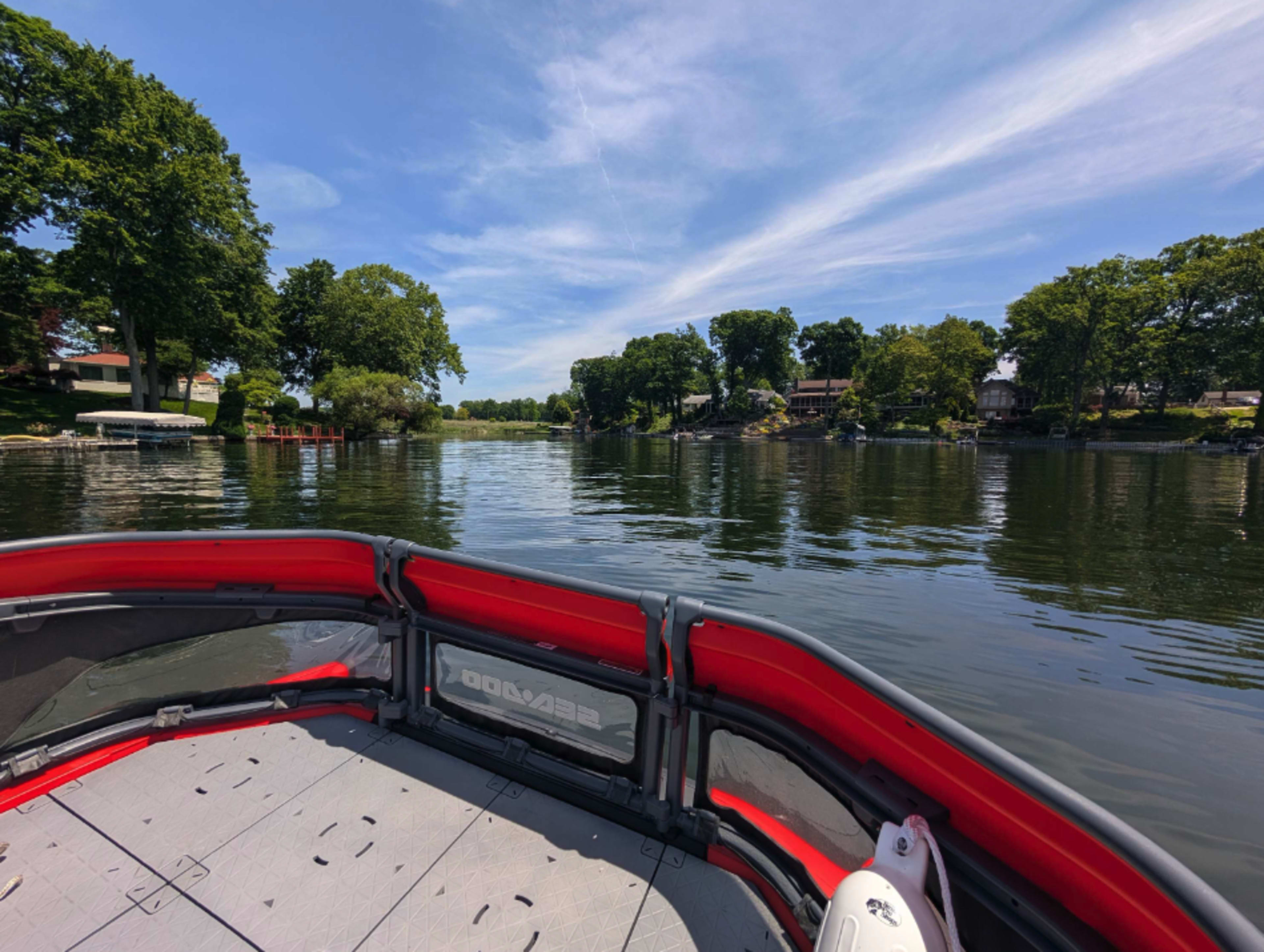 The image shows a view from the back of a boat on a calm lake, with trees lining the shore and houses visible in the distance.