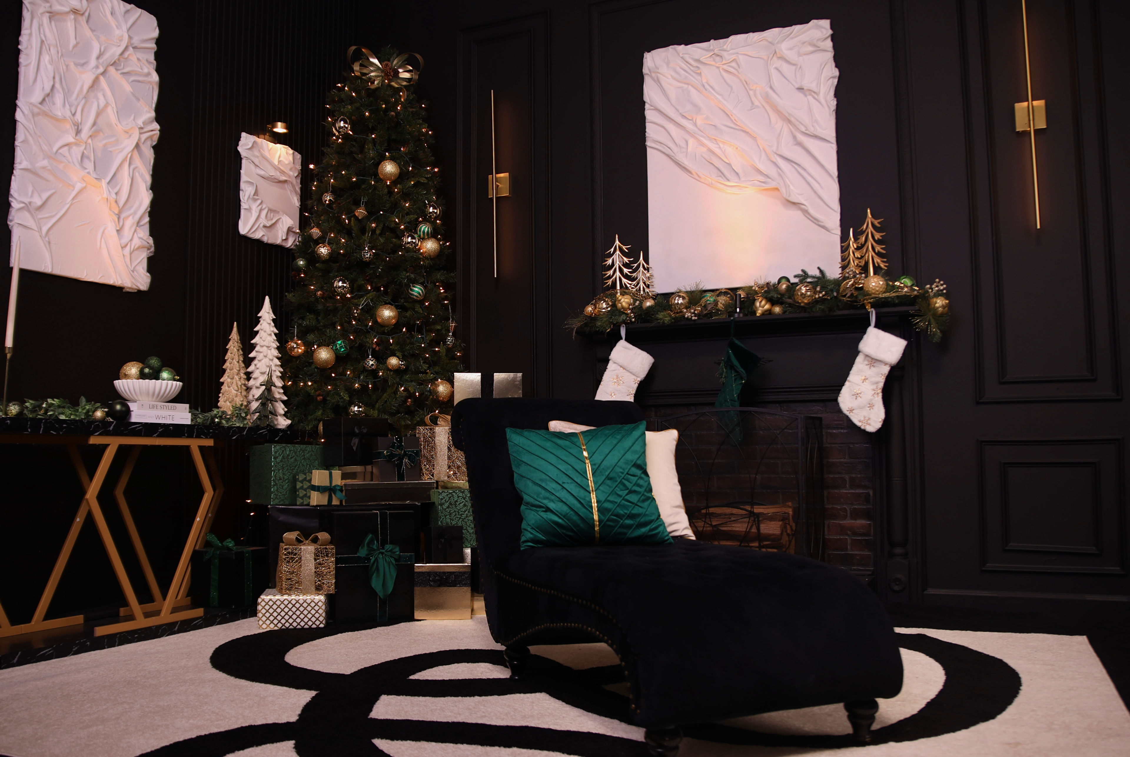 The image shows a stylish holiday-themed living room with a black chaise lounge, a decorated Christmas tree, and festive stockings hanging on the mantel.