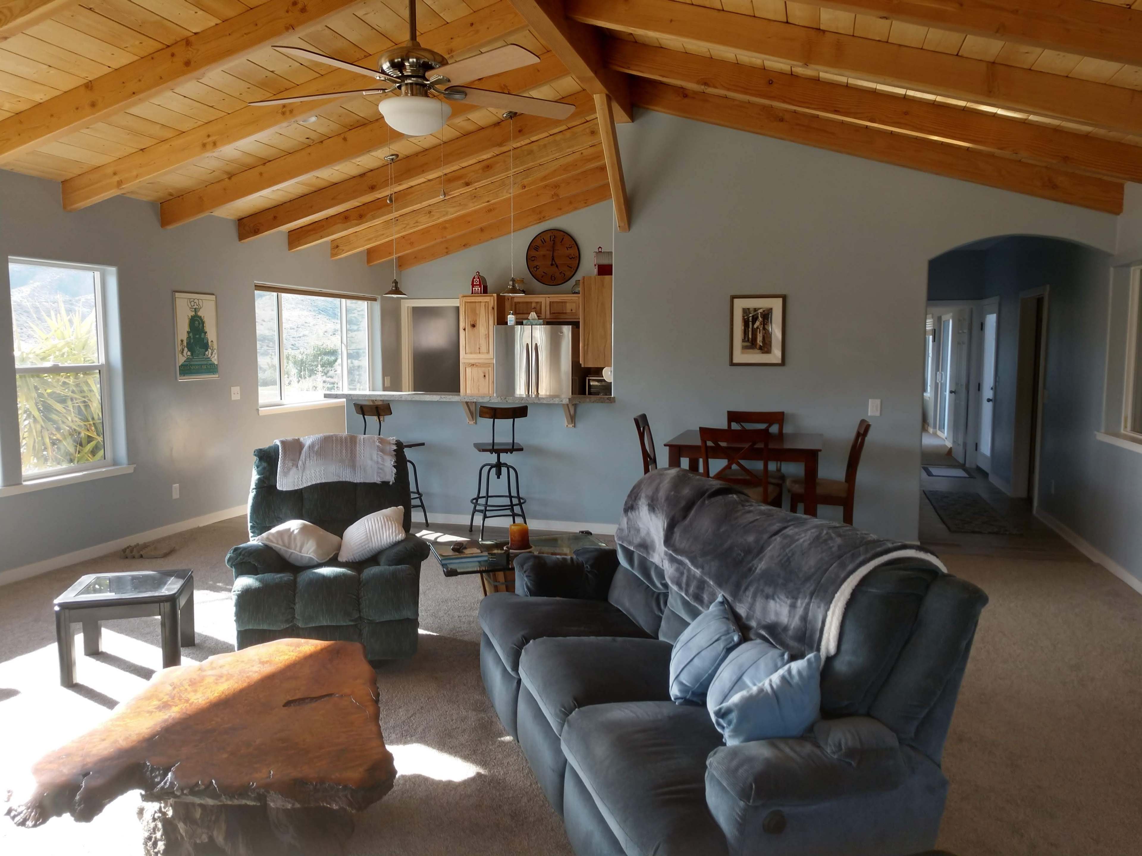 The image shows a cozy living room with a vaulted wooden ceiling, featuring a teal loveseat and armchair, a wooden coffee table, and a dining area with a table and chairs in the background.