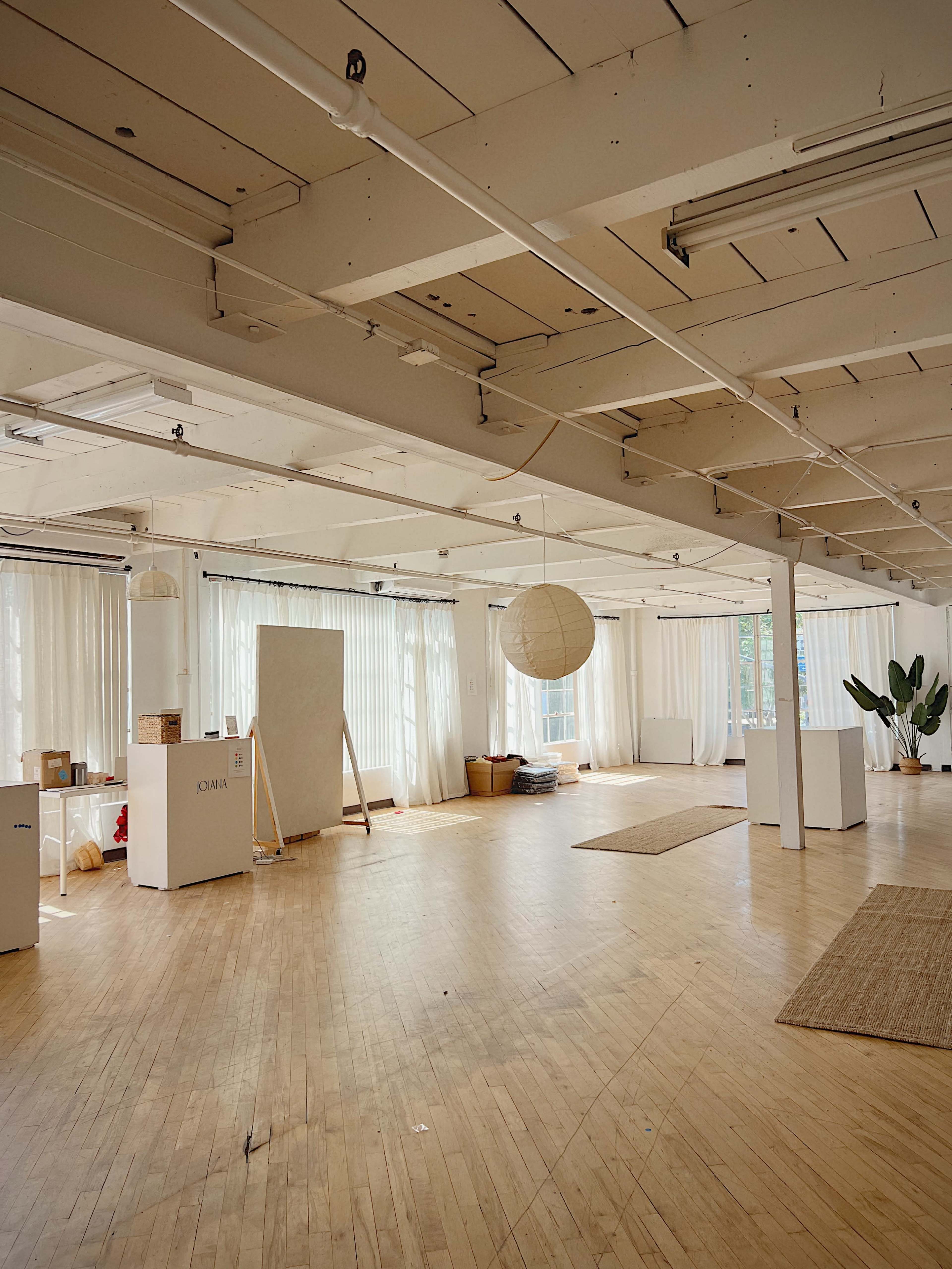 The image shows a spacious, well-lit interior of an empty studio with wooden flooring, large windows, and minimalistic decor.