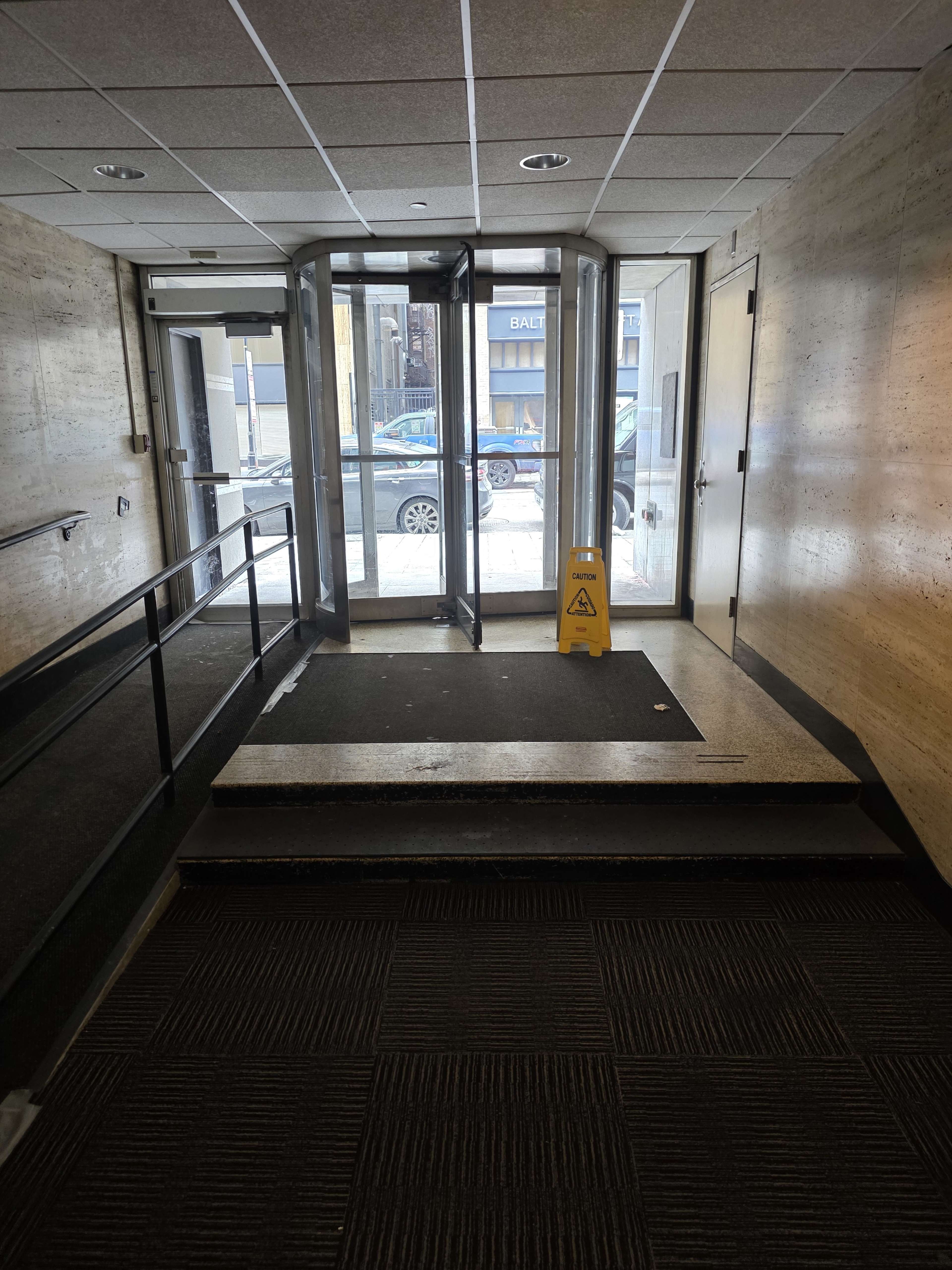 The image shows a brightly lit entrance area with a revolving door, a warning sign placed on the floor, and a sloped pathway leading to the outside.