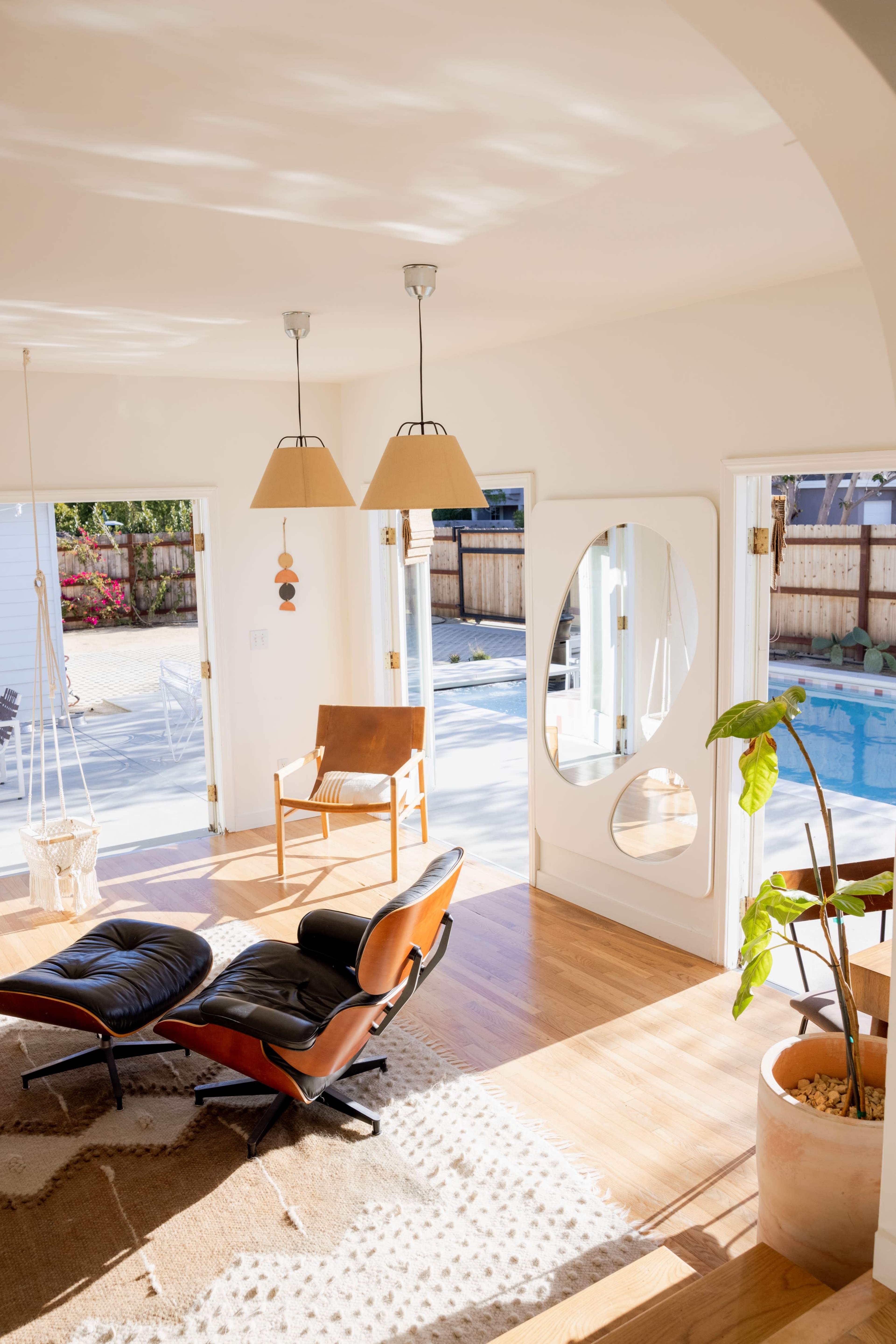 A bright living room features a stylish chair, a large mirror, and open doors leading to a pool area.
