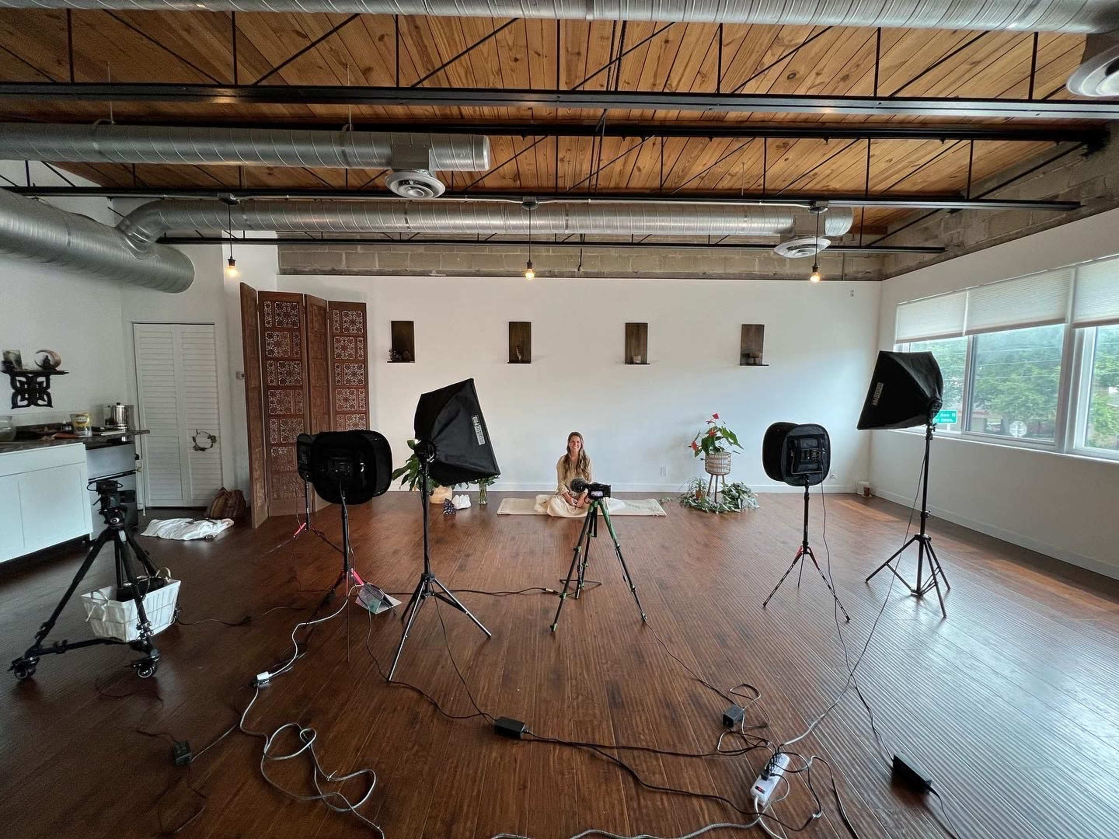 A person sits in a sparsely furnished studio surrounded by photography equipment and overhead lights.