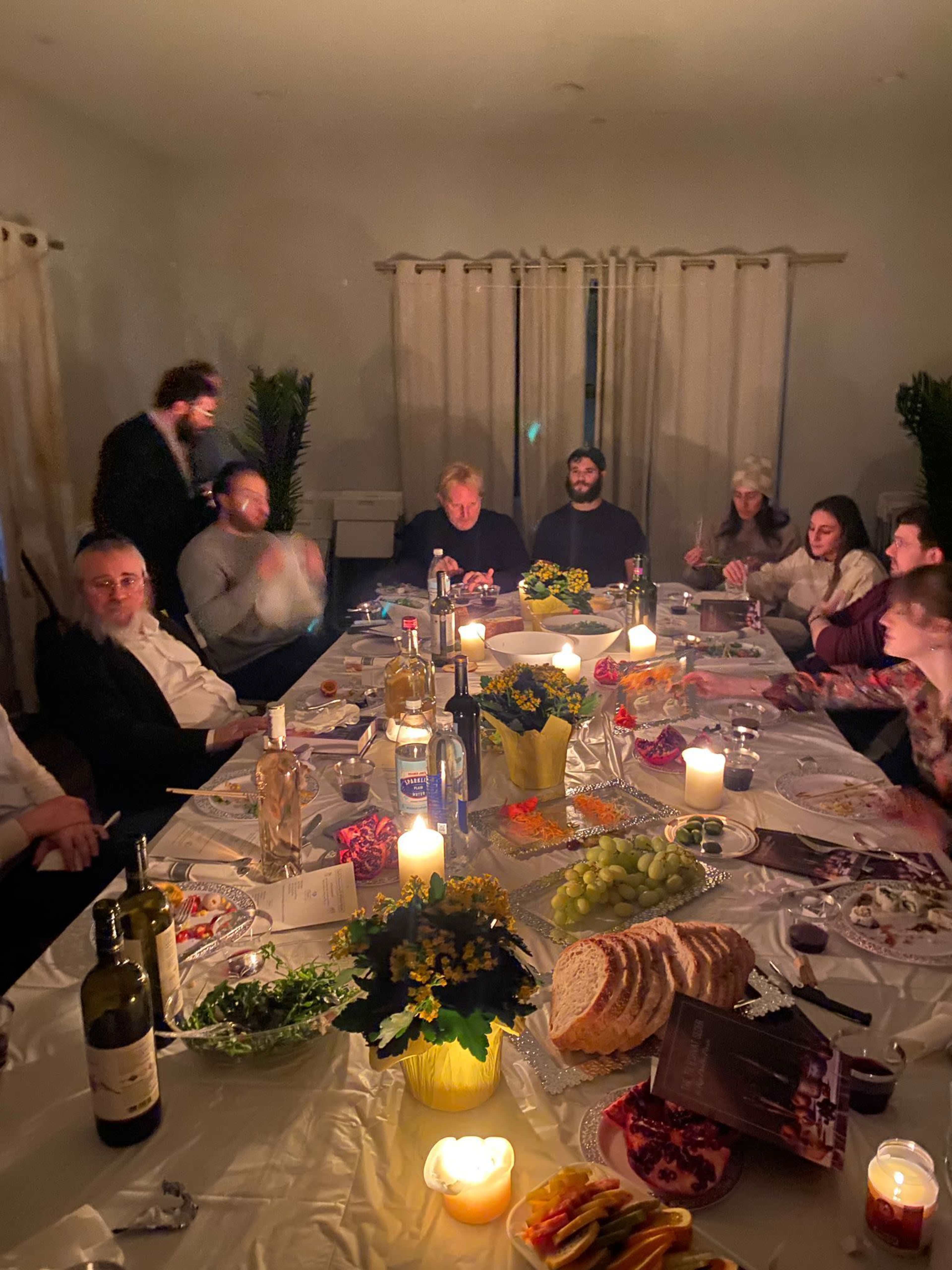 A group of people gathered around a long table covered with food, drinks, and candles in a warmly lit room.