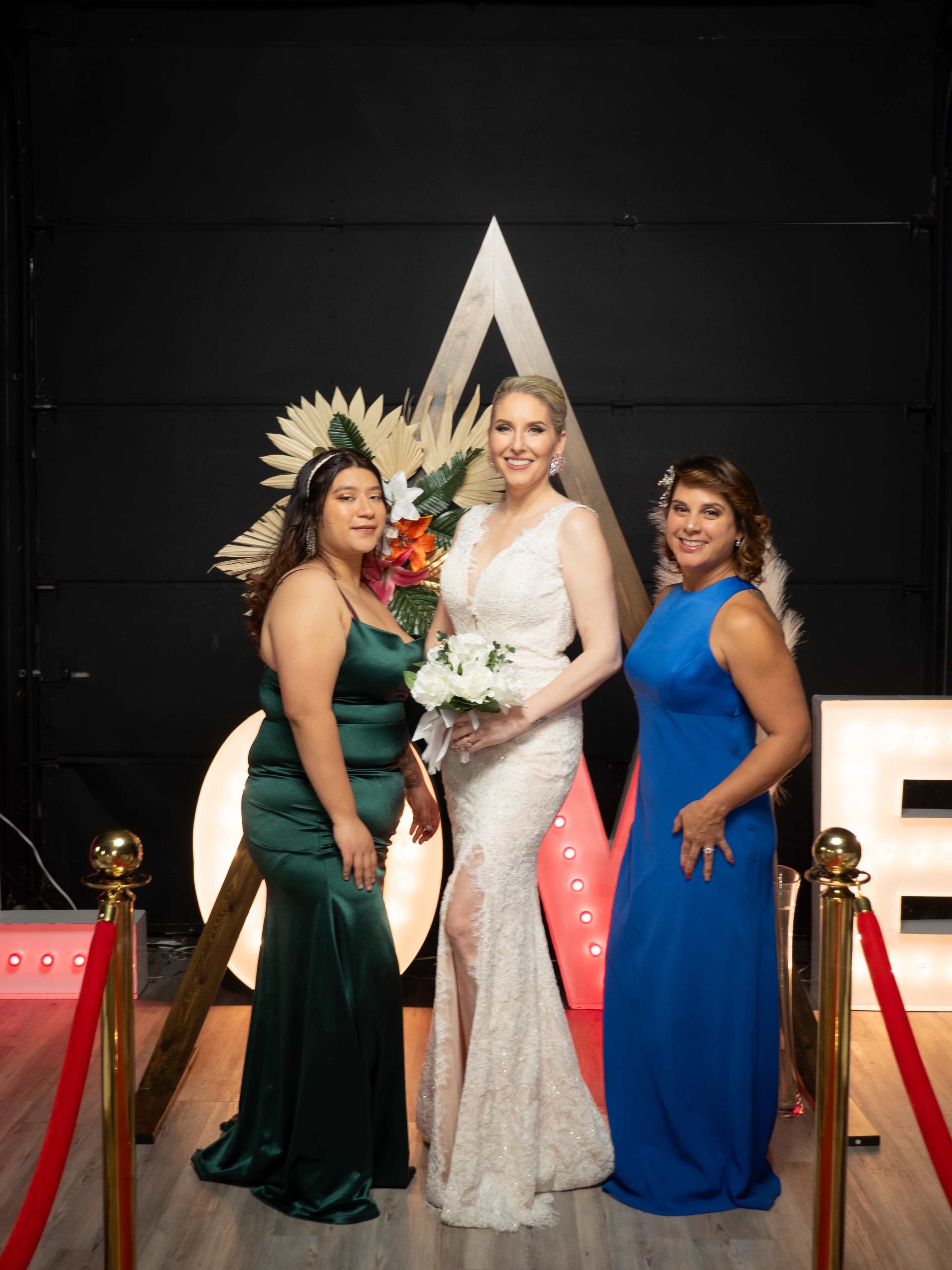 Three women pose together for a photo in front of a decorative backdrop featuring floral arrangements and illuminated letters.