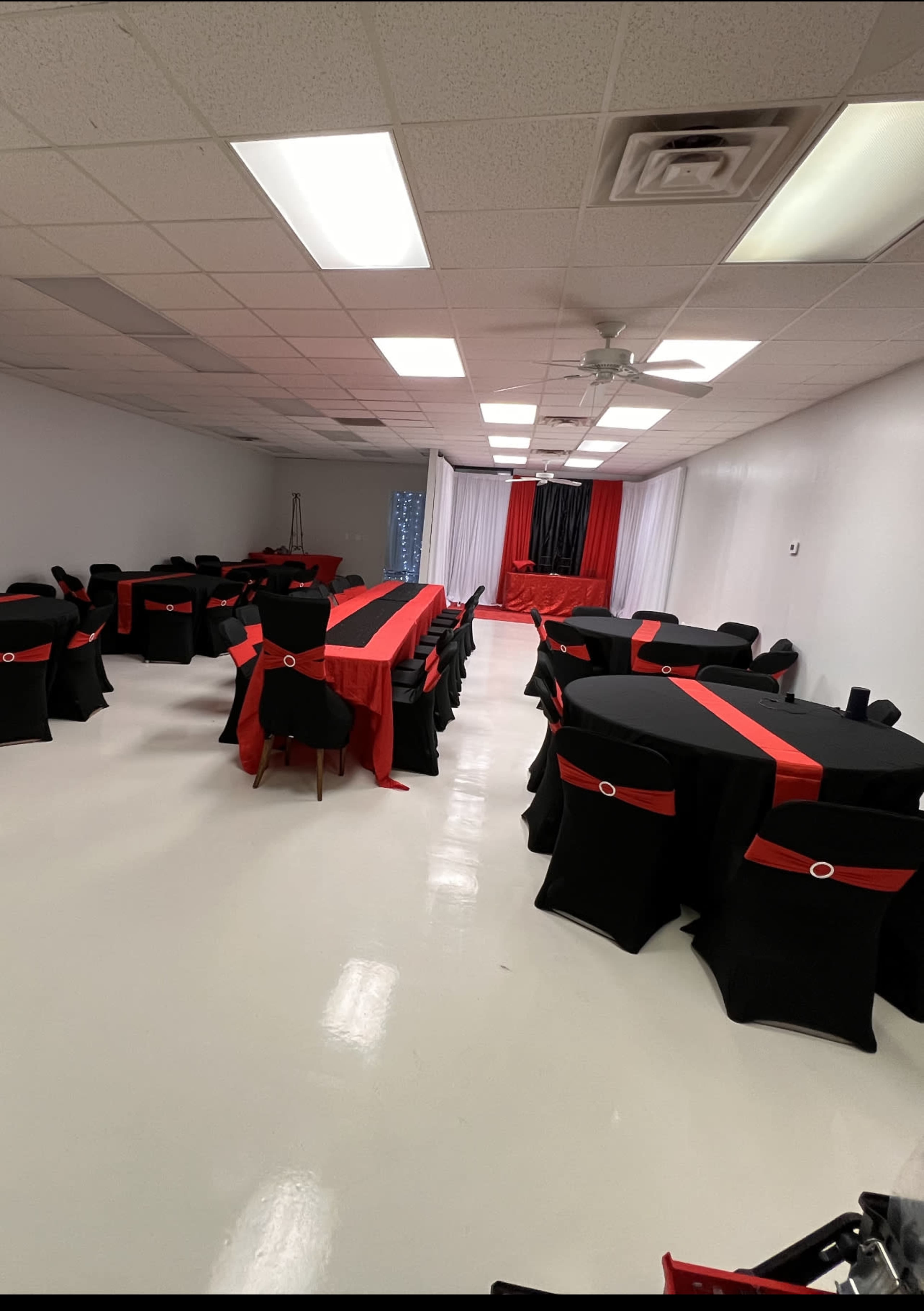 A decorated event space with several round tables covered in black tablecloths featuring red accents, arranged in a spacious, brightly lit room.
