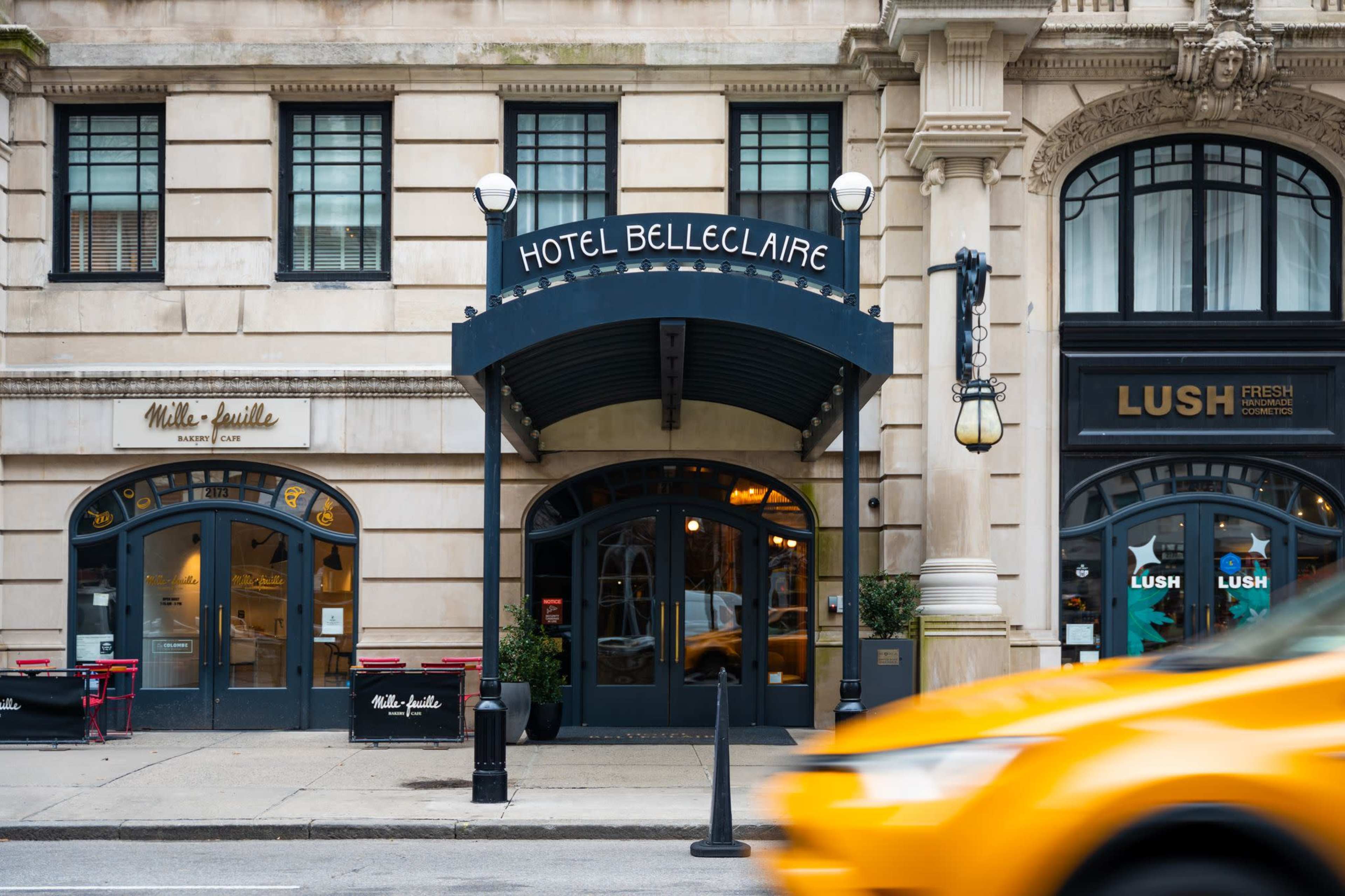 The image shows the entrance of Hotel Belleclaire, flanked by a cafe and a Lush store, with a yellow taxi passing by on the street.