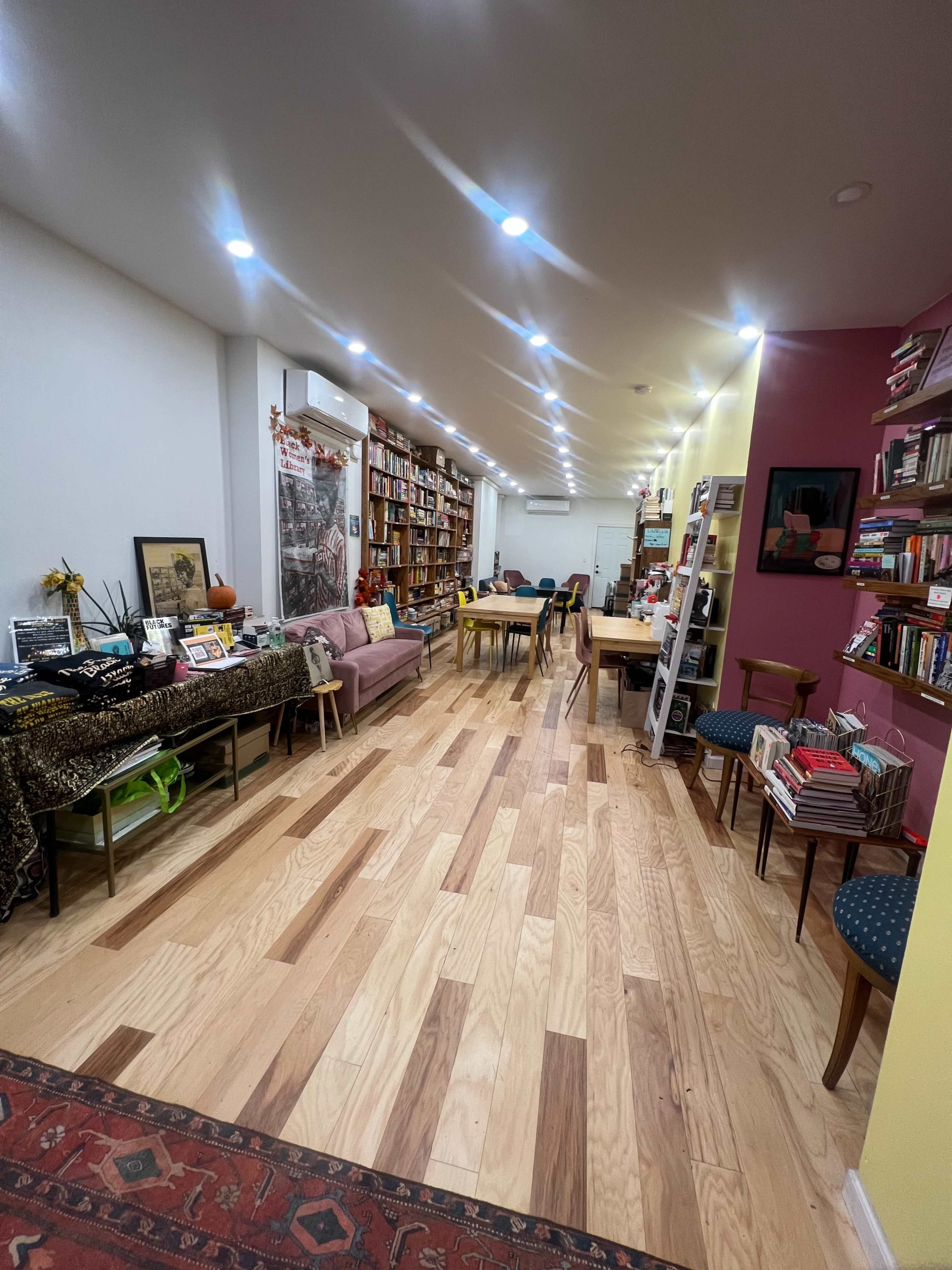A well-lit room filled with bookshelves, a pink couch, and various tables arranged along a wooden floor.