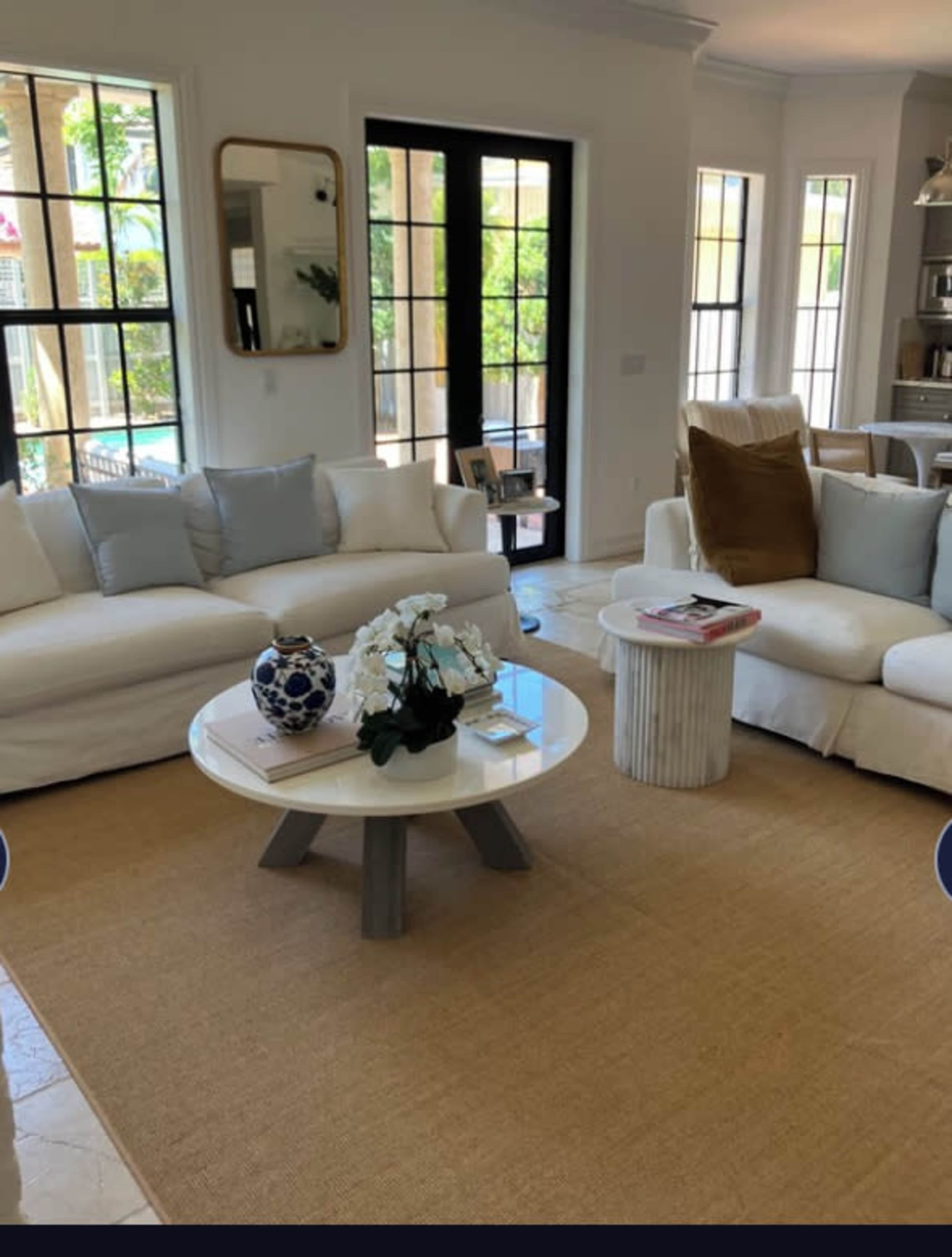 The image shows a living room with two white sofas, a round coffee table, and a neutral-colored rug, surrounded by large windows.