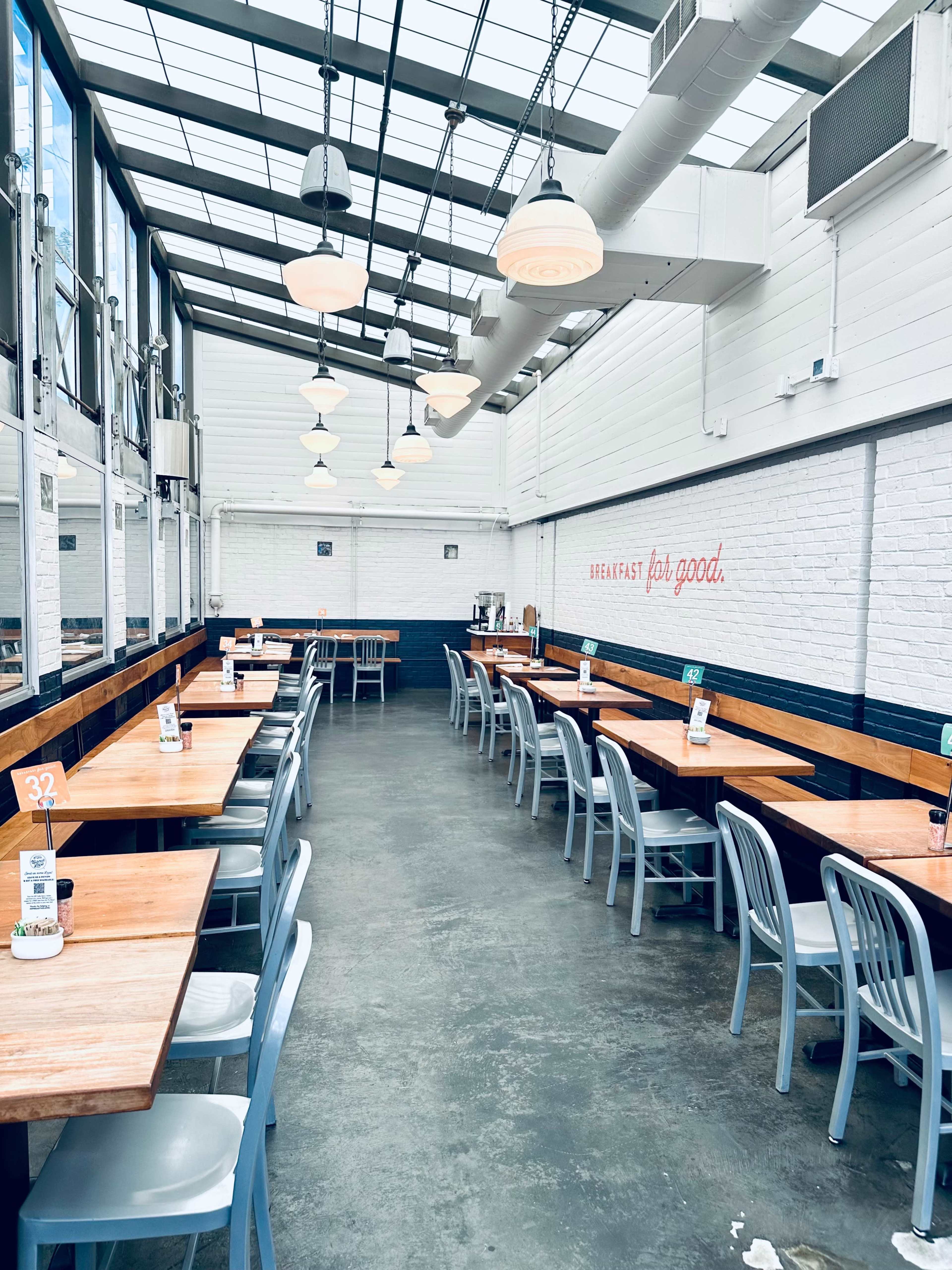 The image shows a bright, spacious restaurant interior with wooden tables, gray chairs, and a large skylight overhead.