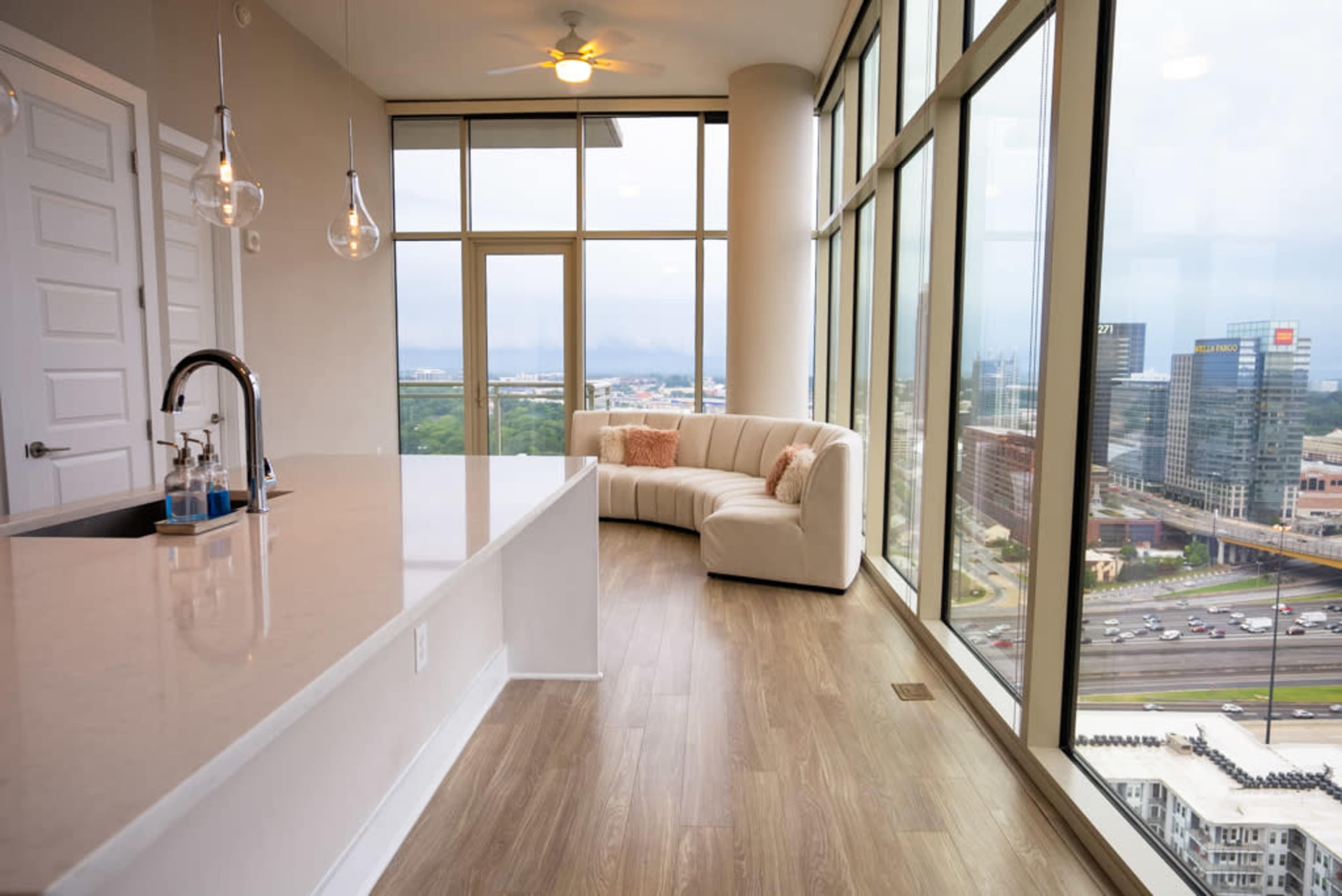 The image shows a modern apartment interior with a curved white couch and large floor-to-ceiling windows overlooking a cityscape.