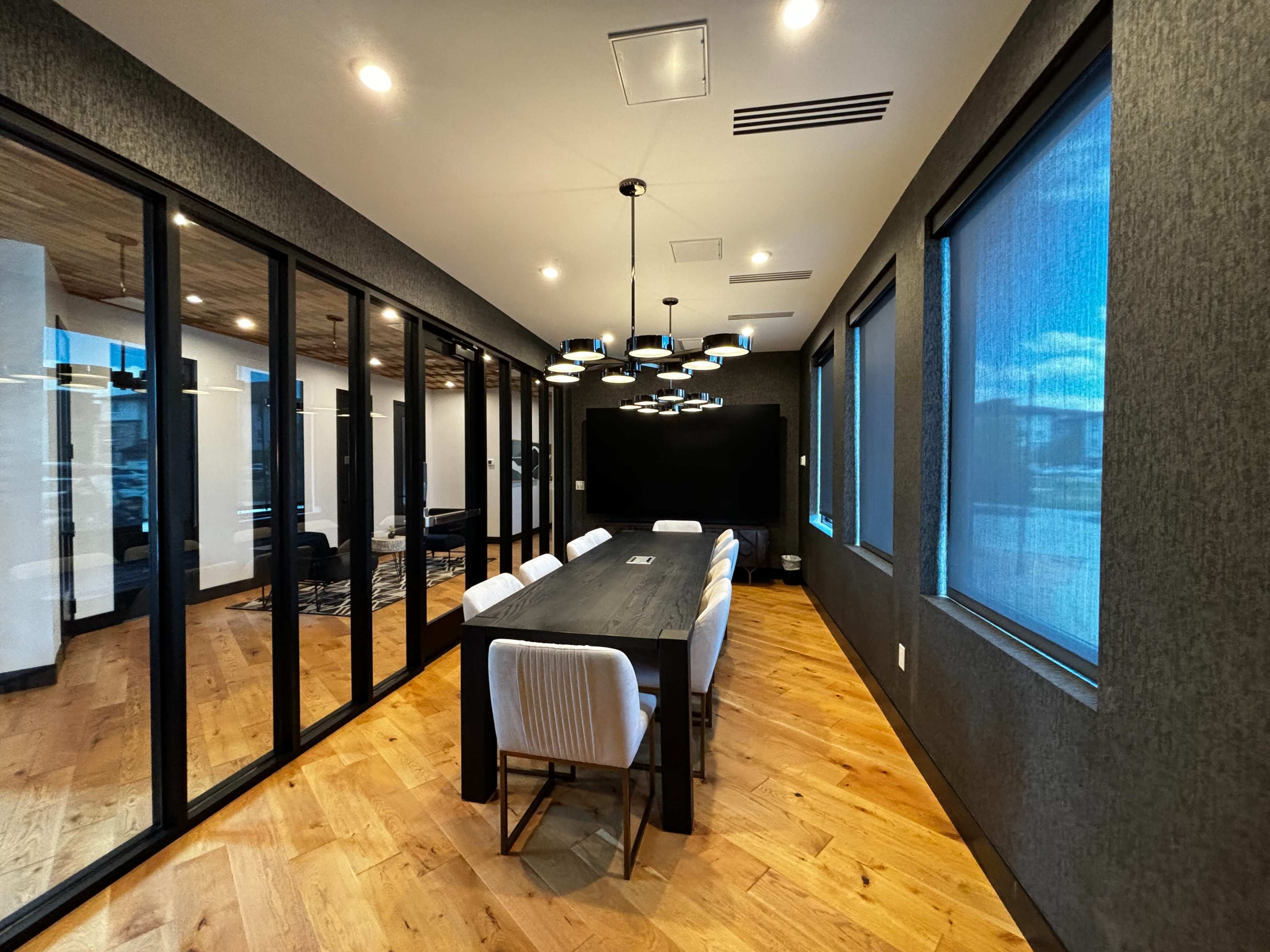 A modern conference room features a long table surrounded by chairs, with glass walls and large windows allowing natural light to enter.