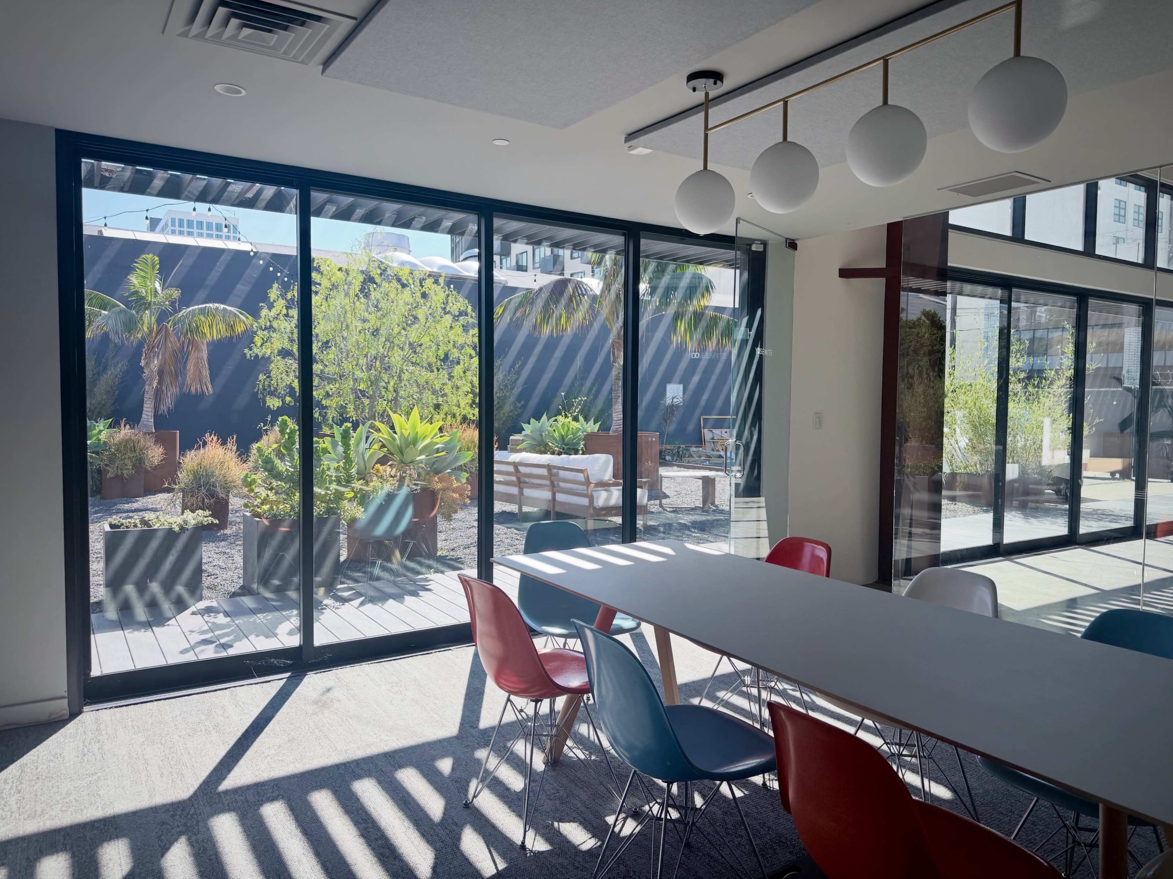A modern meeting room features a long table surrounded by colorful chairs, with large glass doors opening to an outdoor garden area filled with plants.