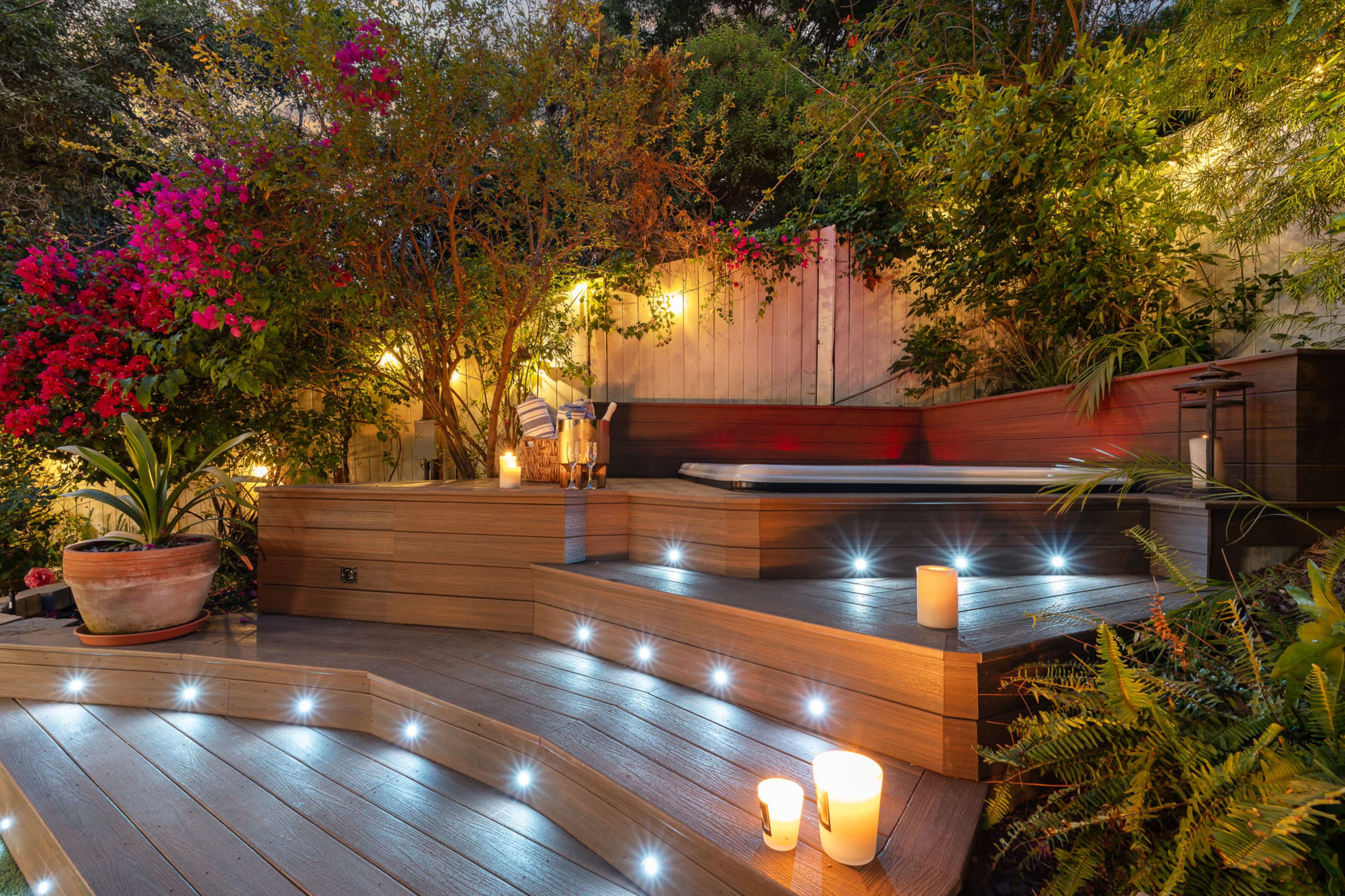 A landscaped outdoor area featuring a hot tub, illuminated steps with LED lights, and lush greenery surrounded by blooming flowers.