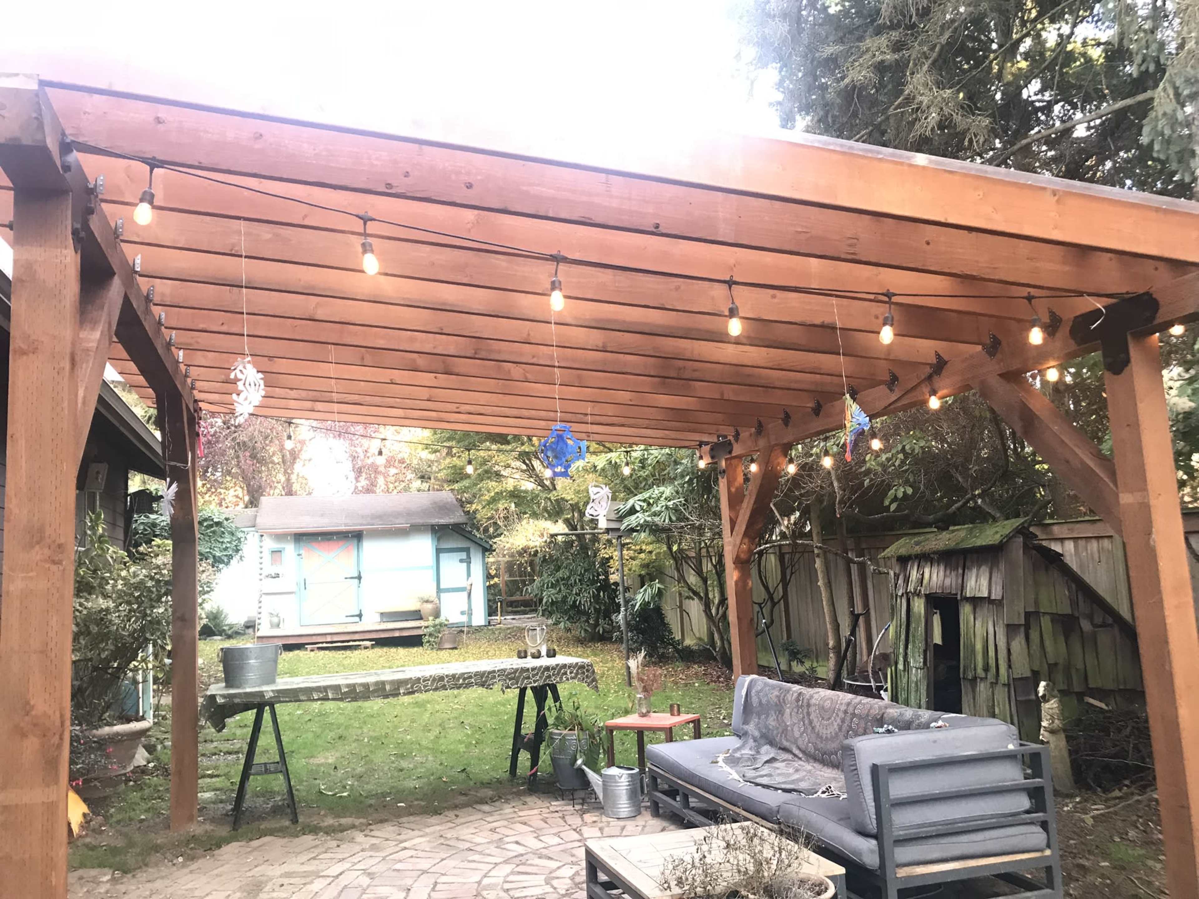 A wooden pergola with hanging lights overlooks a grassy backyard where a covered table and a seating area are arranged.