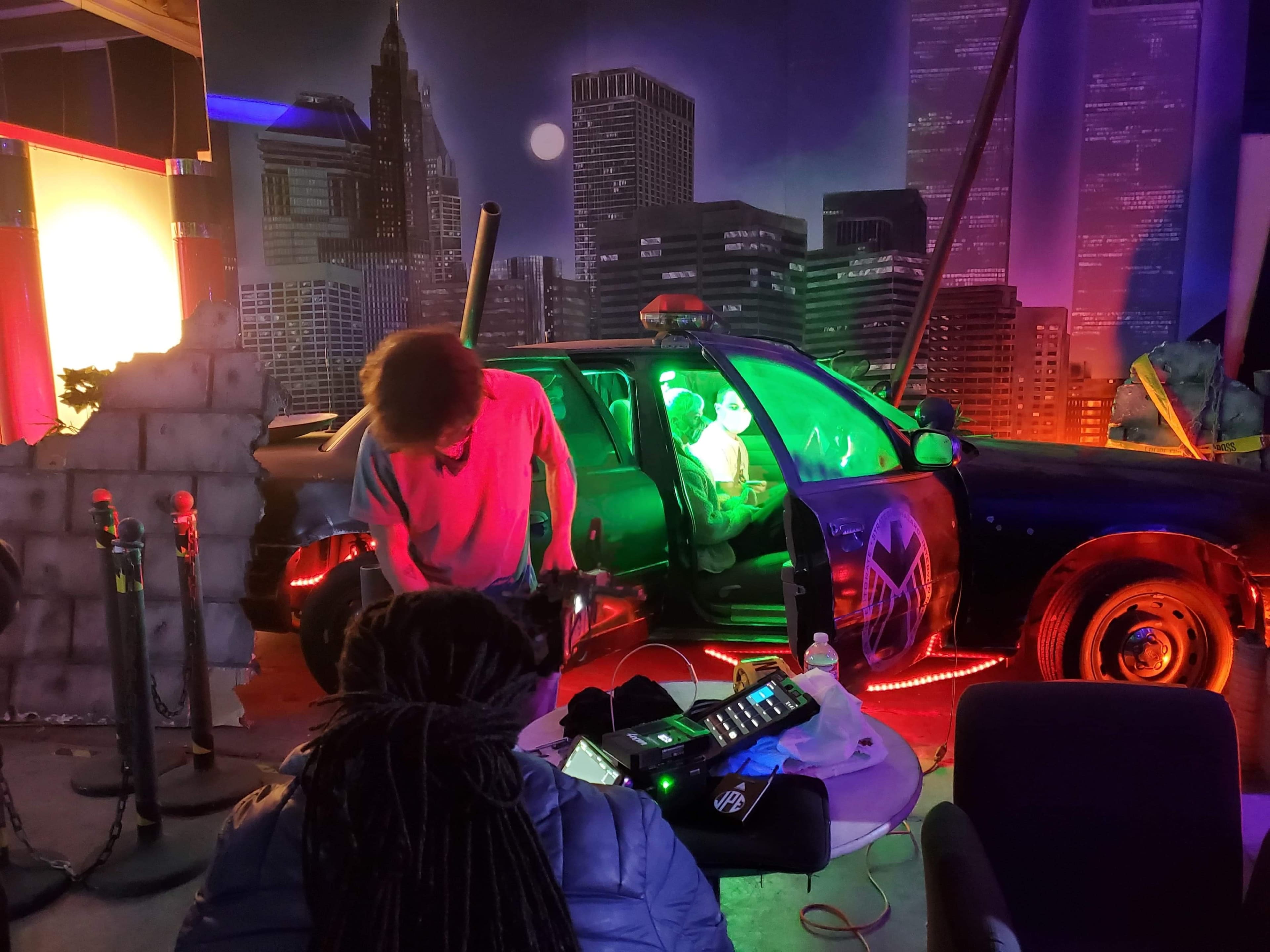 A person stands beside a police car illuminated with green lights, while another individual sits inside the car amidst a staged urban backdrop.