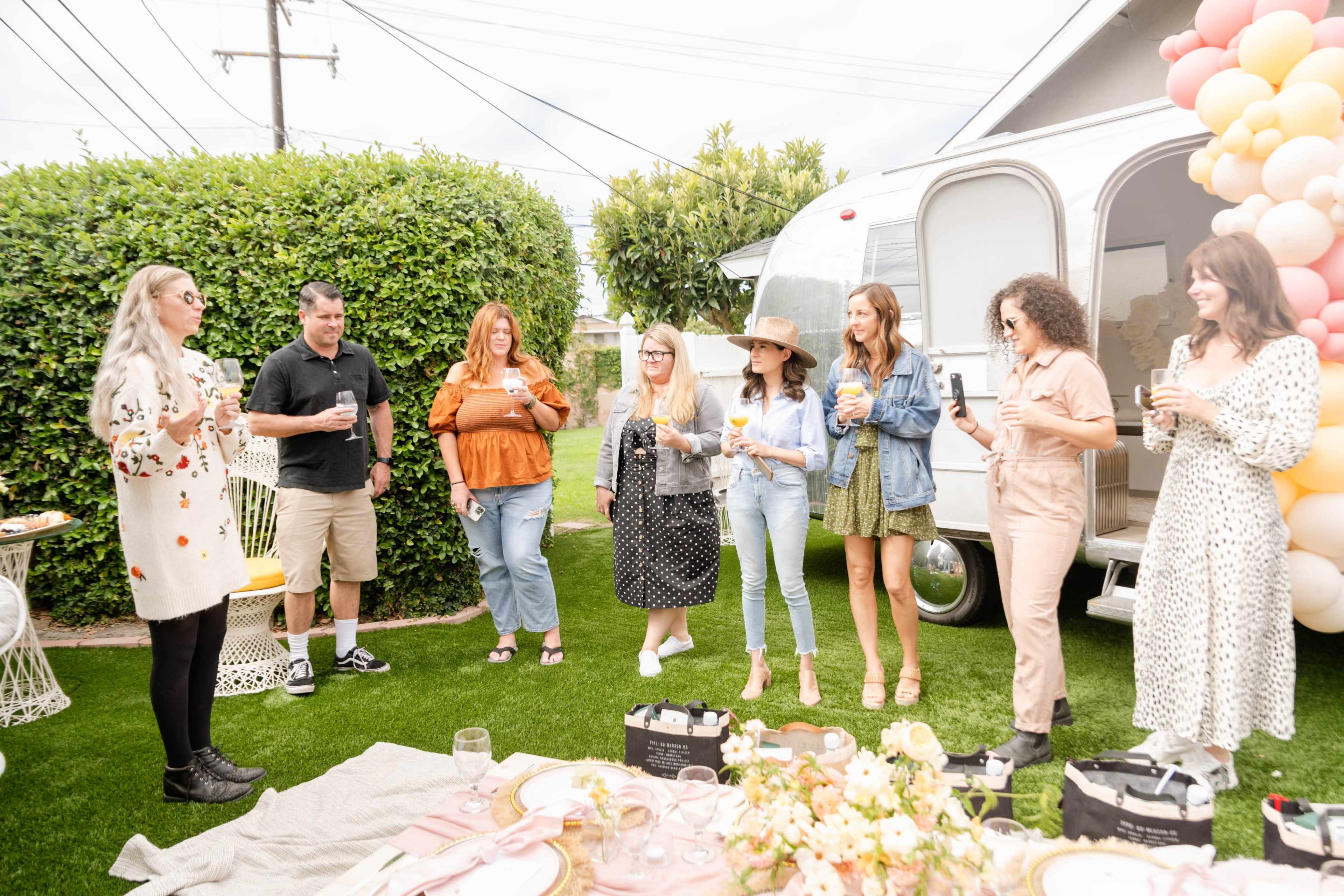 A group of eight women and one man stands in a backyard, holding drinks and gathered around a decorative table near a silver trailer and colorful balloon arch.