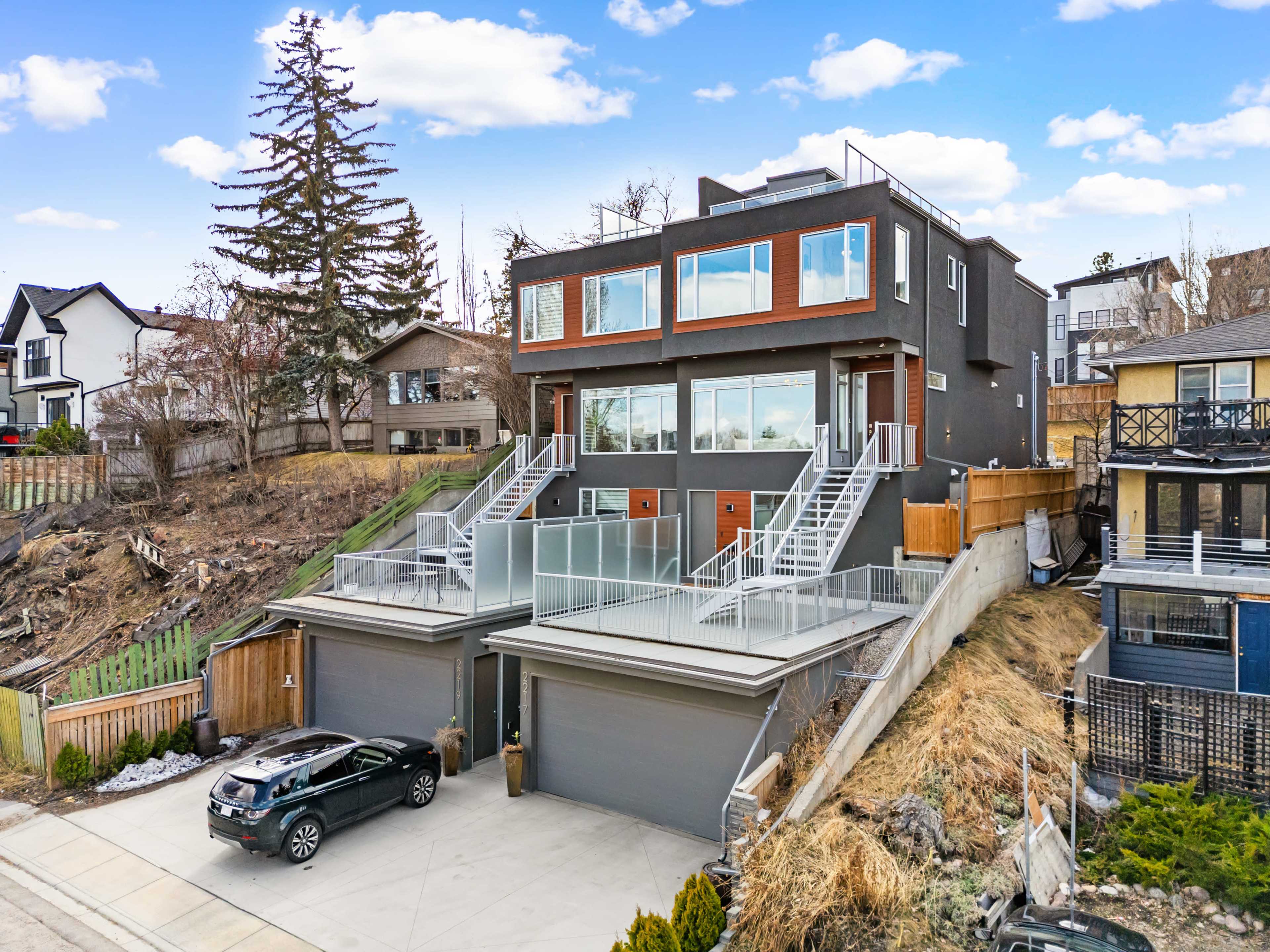 The image shows a modern duplex with large windows, two separate entries, and balconies on a hillside, surrounded by trees and residential properties.