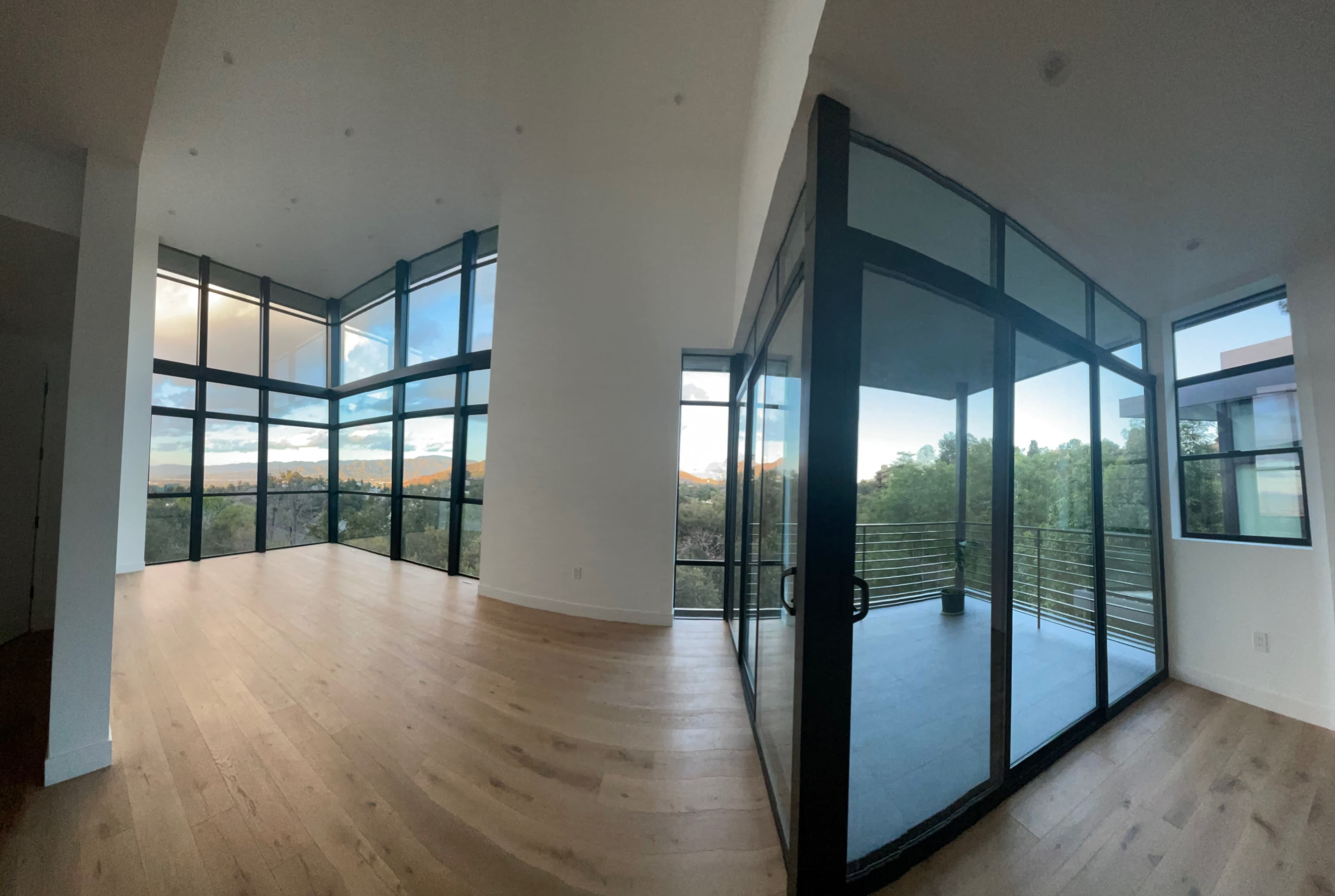 The image shows a modern, spacious room with large glass windows offering views of the outdoors and a wooden floor.
