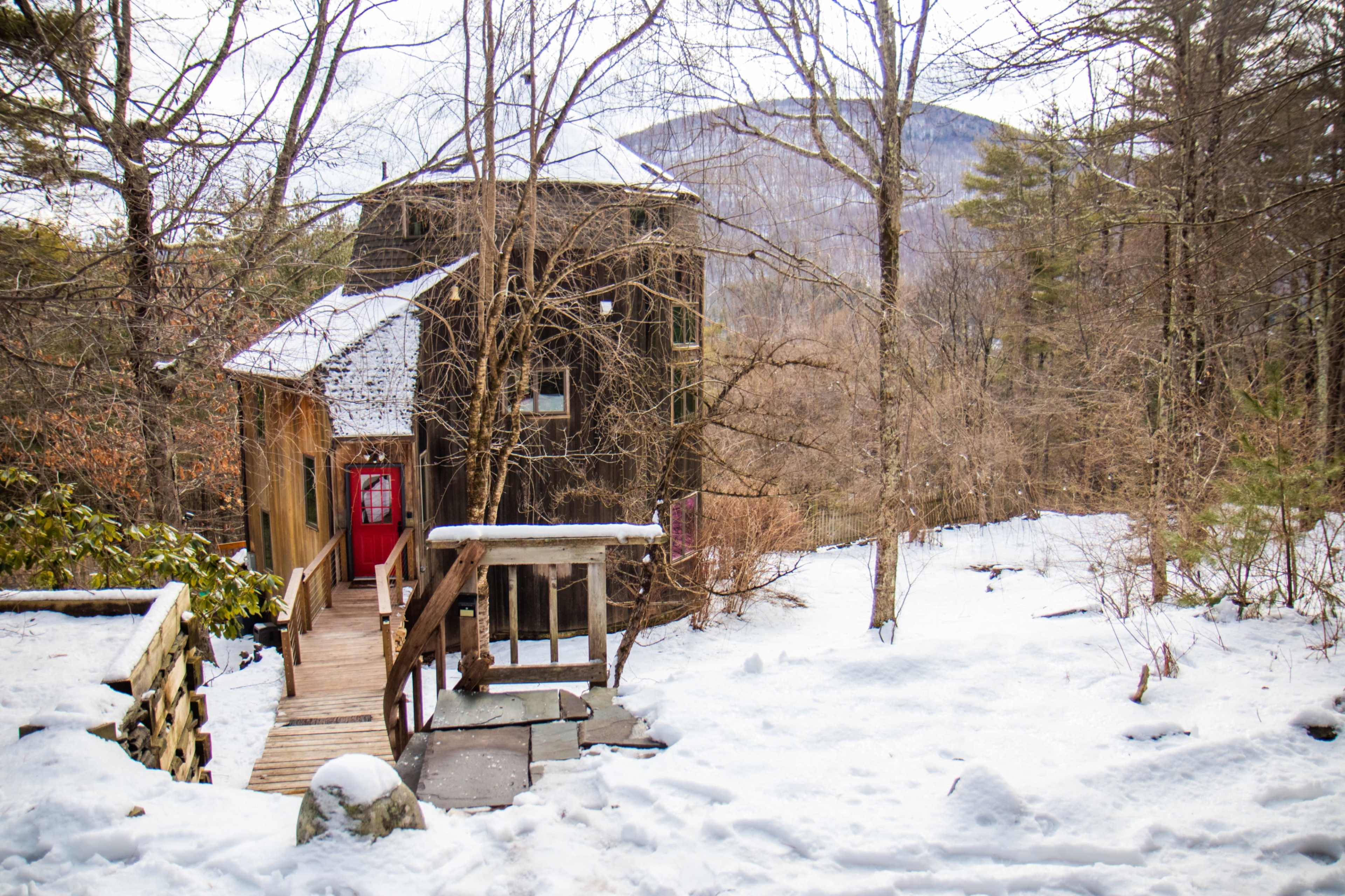 A two-story cabin with a red door and large windows is set against a snowy landscape and forested hillside.
