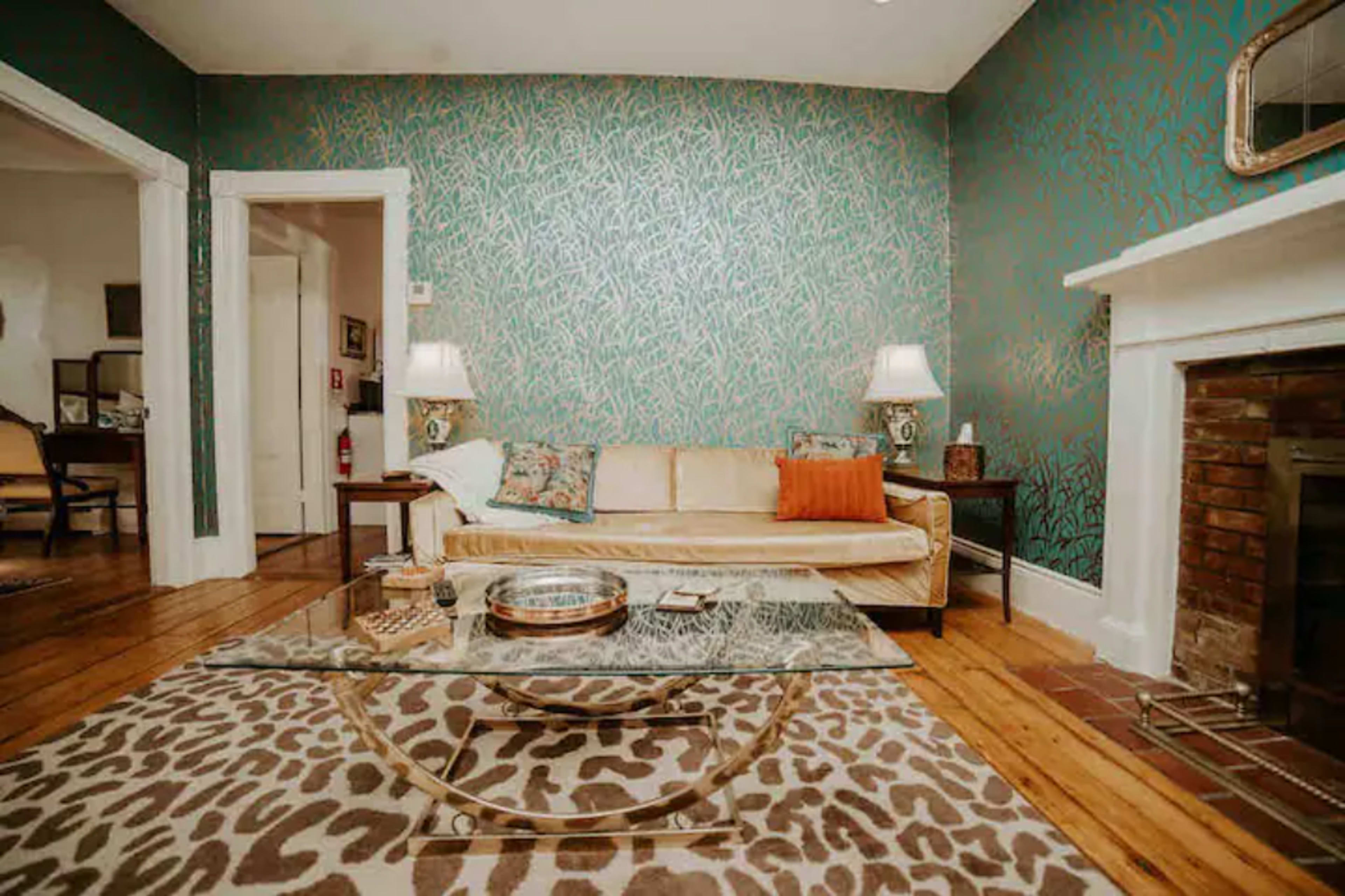 The living room features a patterned wallpaper, a beige sofa with an orange pillow, a glass coffee table, and a fireplace with a brick surround.