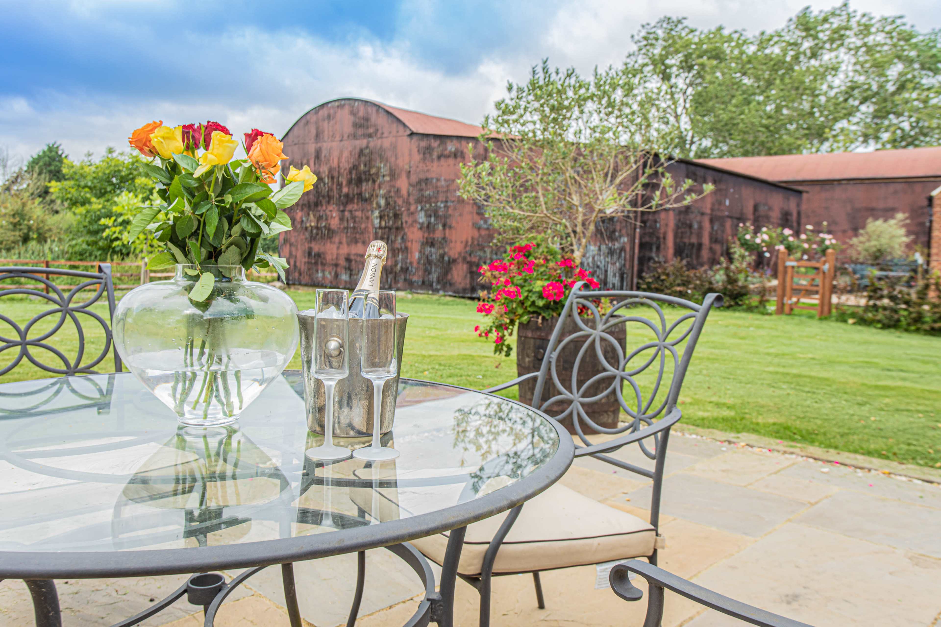 A glass table with a bouquet of roses and a bottle of champagne in an ice bucket is set on a patio overlooking a grassy area and a barn in the background.