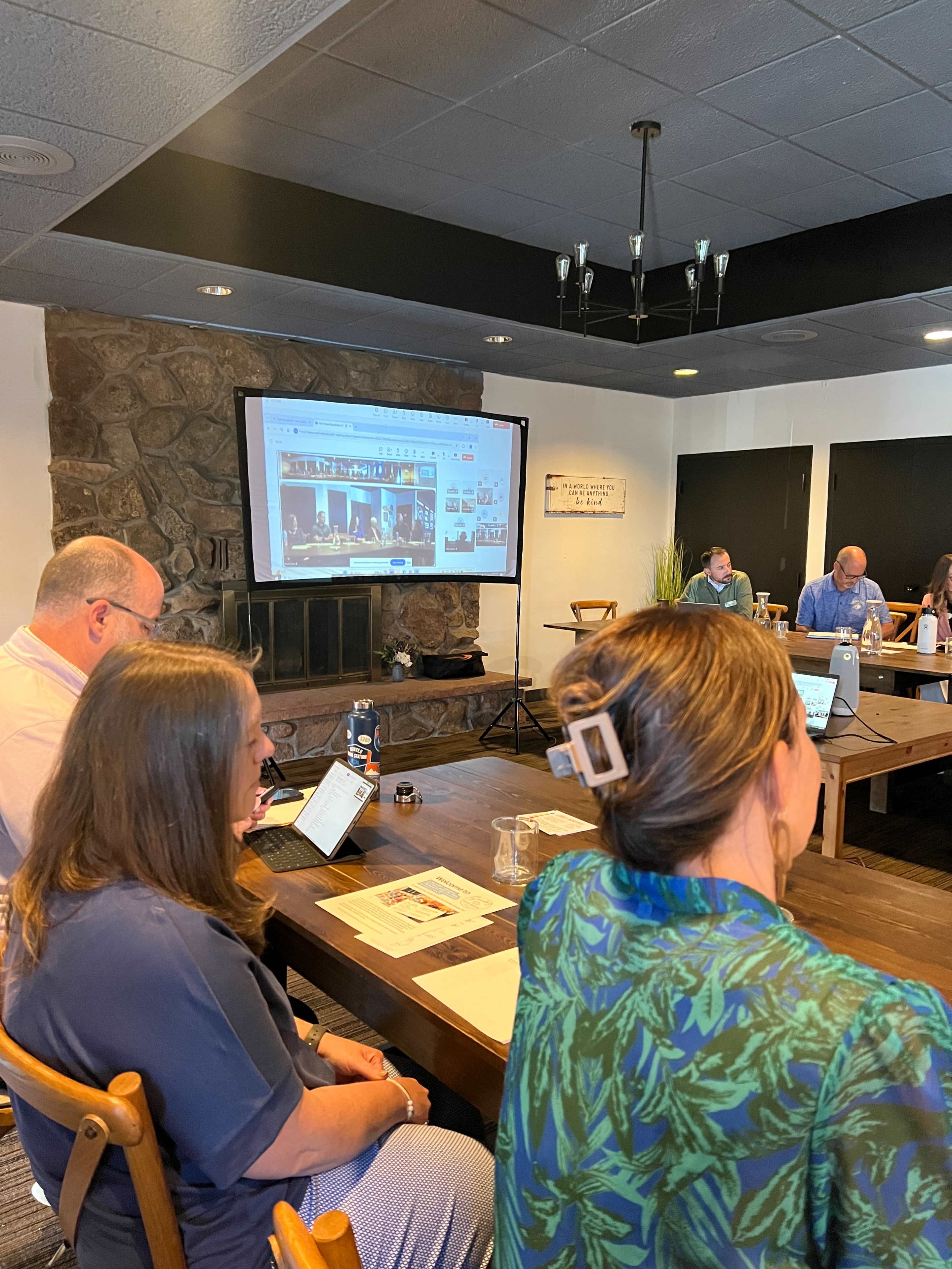 A group of people are seated around a table in a conference room, with a projector screen displaying images in the background.