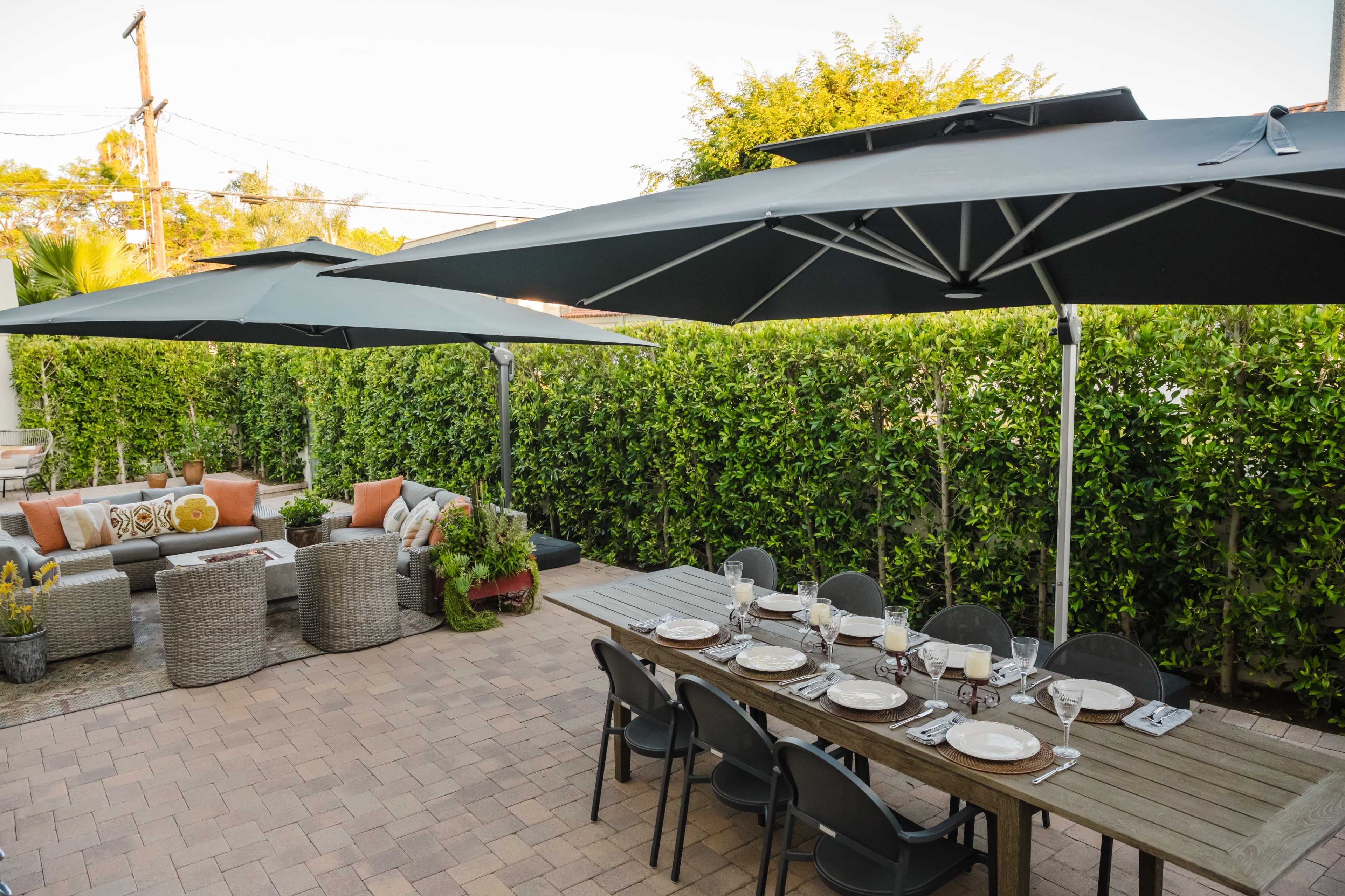 The image shows a backyard patio with a dining table set for a meal, surrounded by various outdoor seating arrangements and large umbrellas.