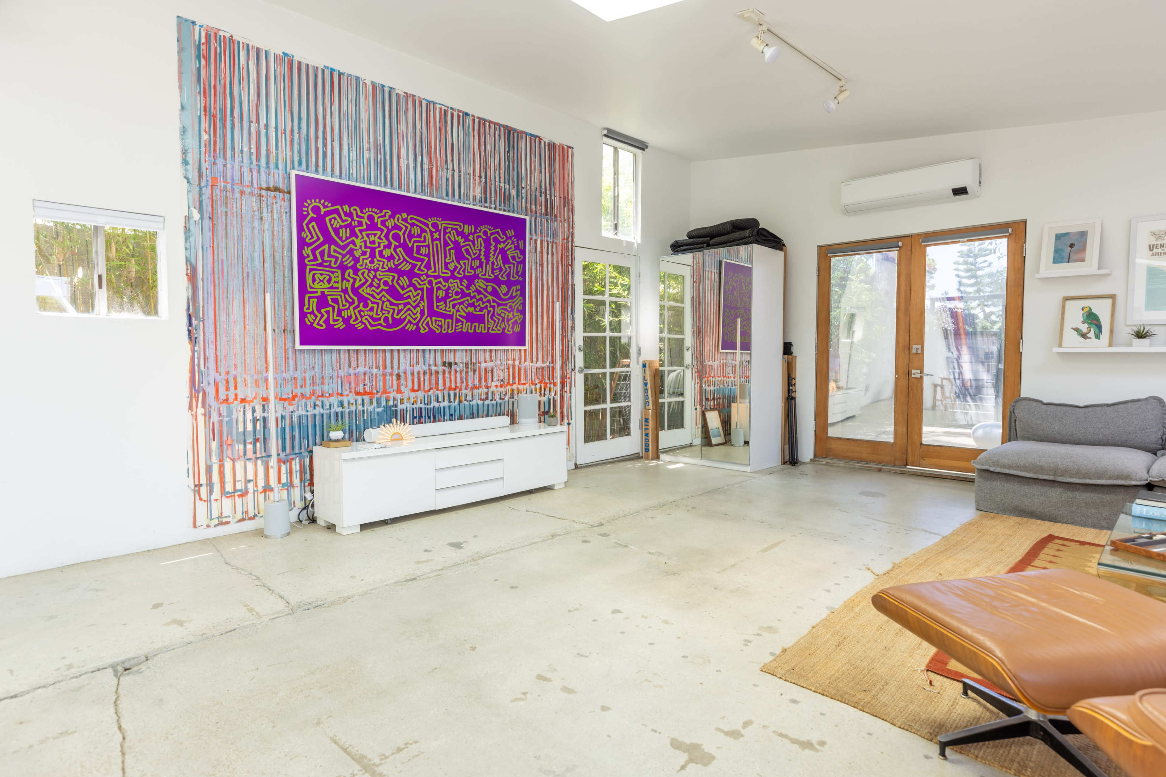 The room features a minimalist design with a large abstract purple artwork on the wall, a gray sofa, and glass doors leading outside.