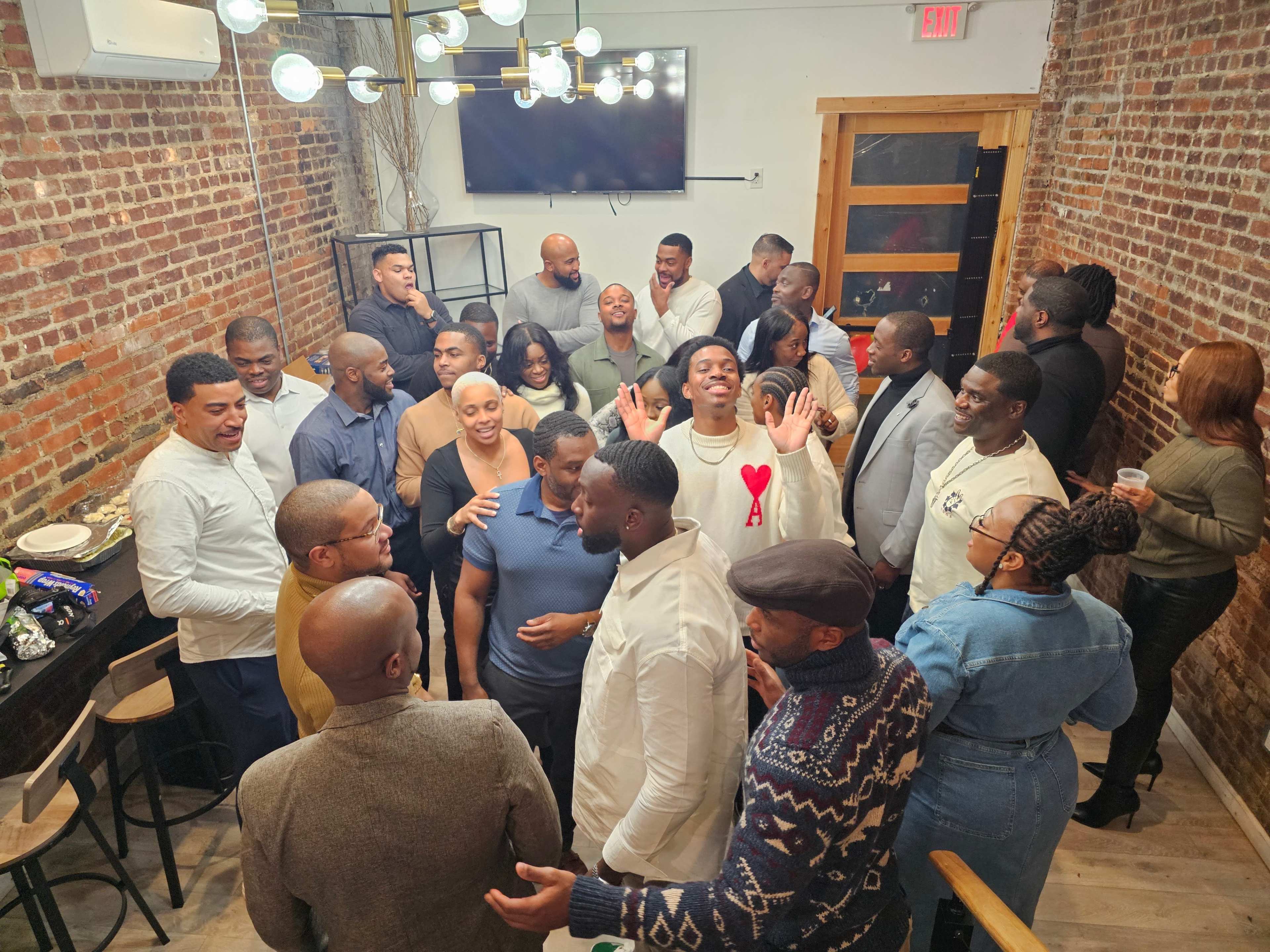 A group of people is socializing in a lively setting with a brick wall, modern lighting, and a mix of expressions and gestures.
