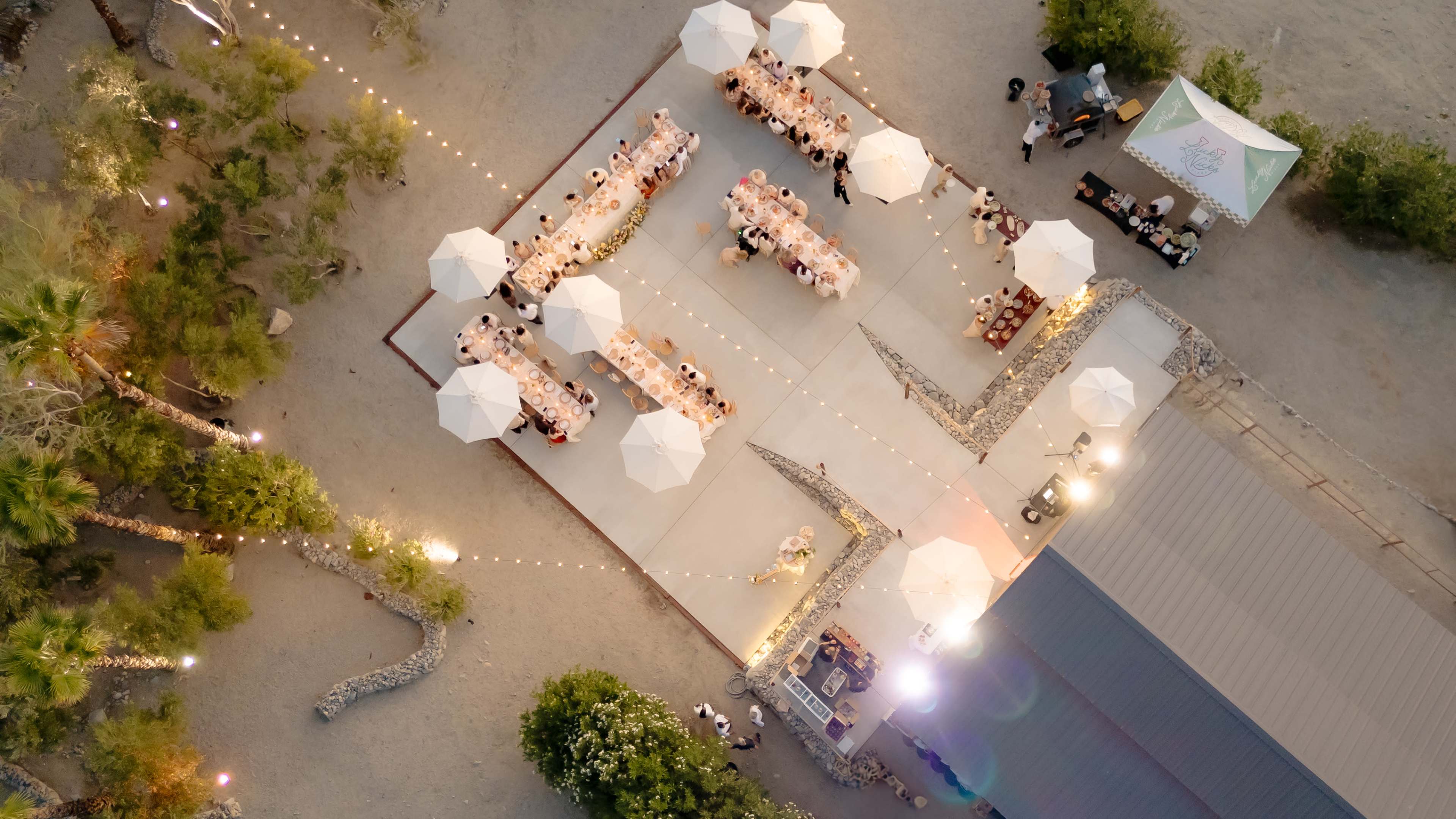An outdoor event features multiple tables set up under umbrellas, arranged in a square formation with string lights illuminating the area.