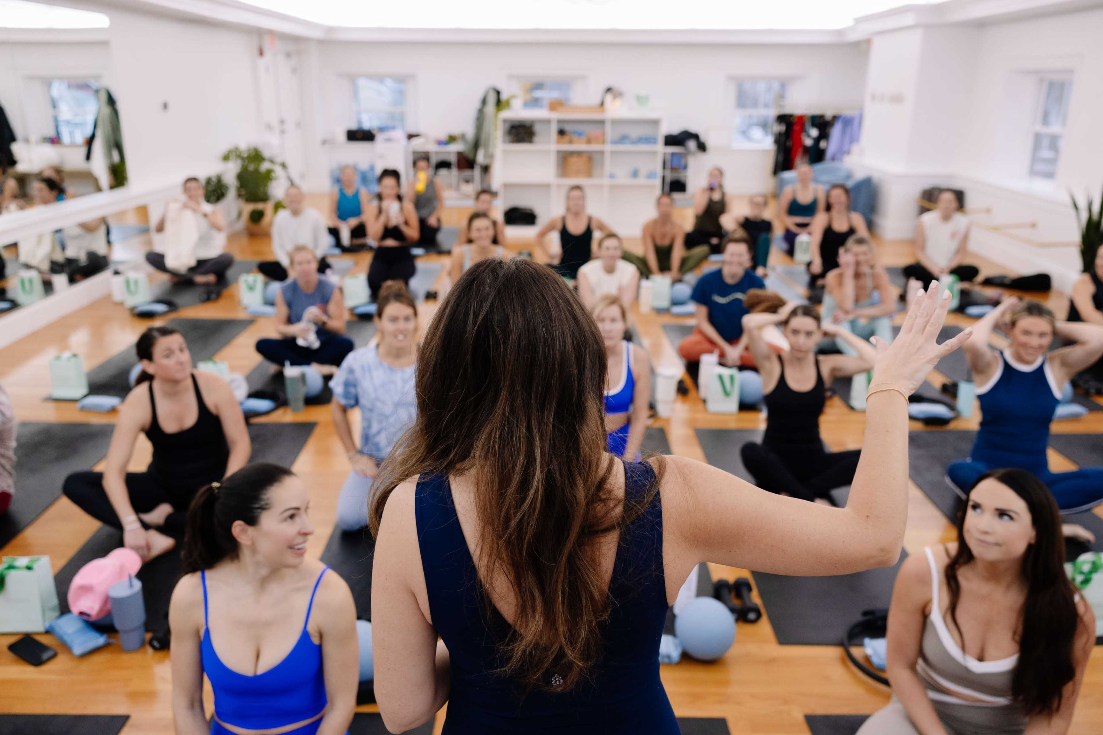 A fitness instructor addresses a group of seated participants in a yoga studio.
