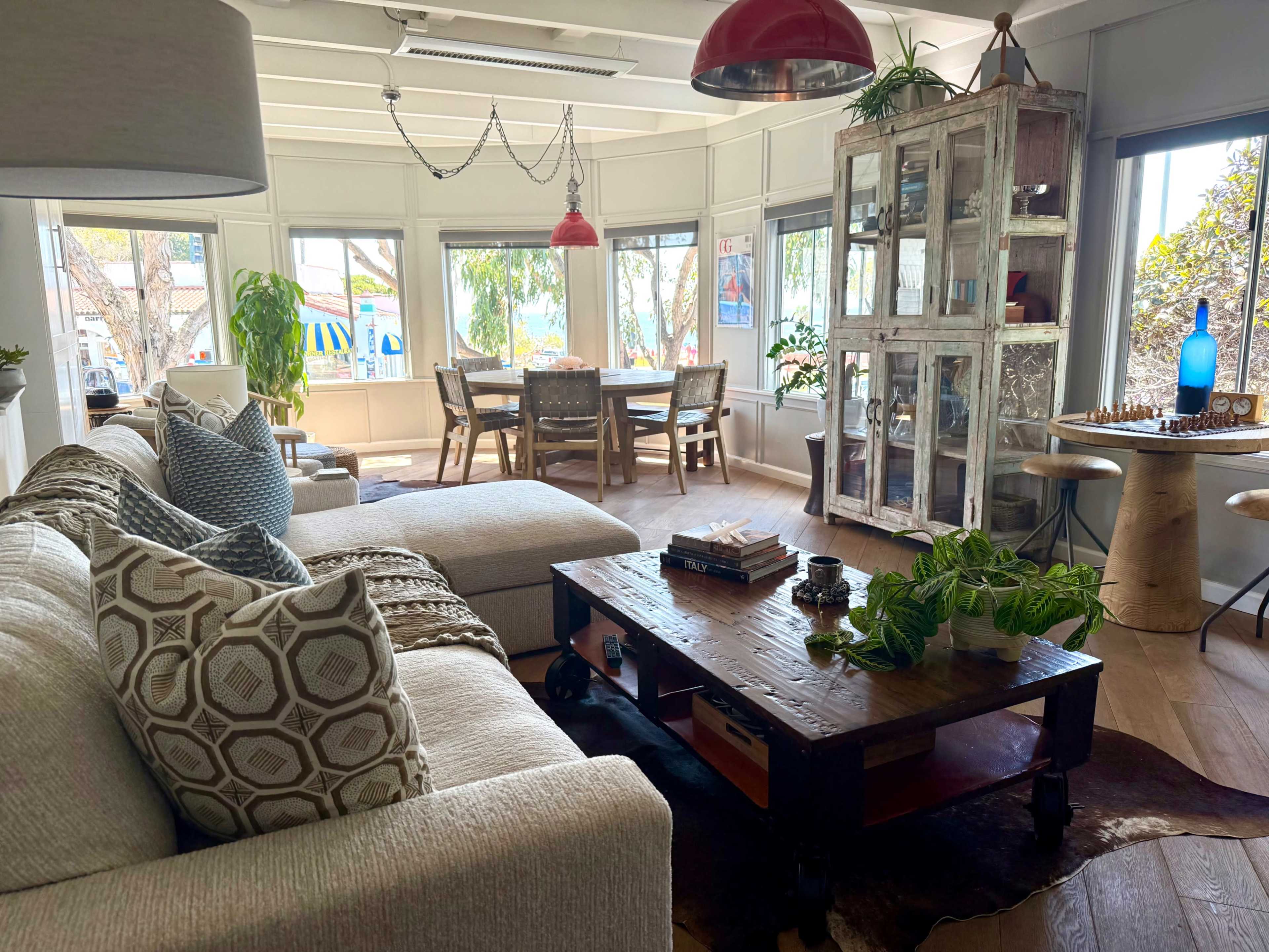 The image shows a bright living room with a large sectional sofa, a wooden coffee table, and a dining area featuring a glass cabinet and a wooden table surrounded by chairs.