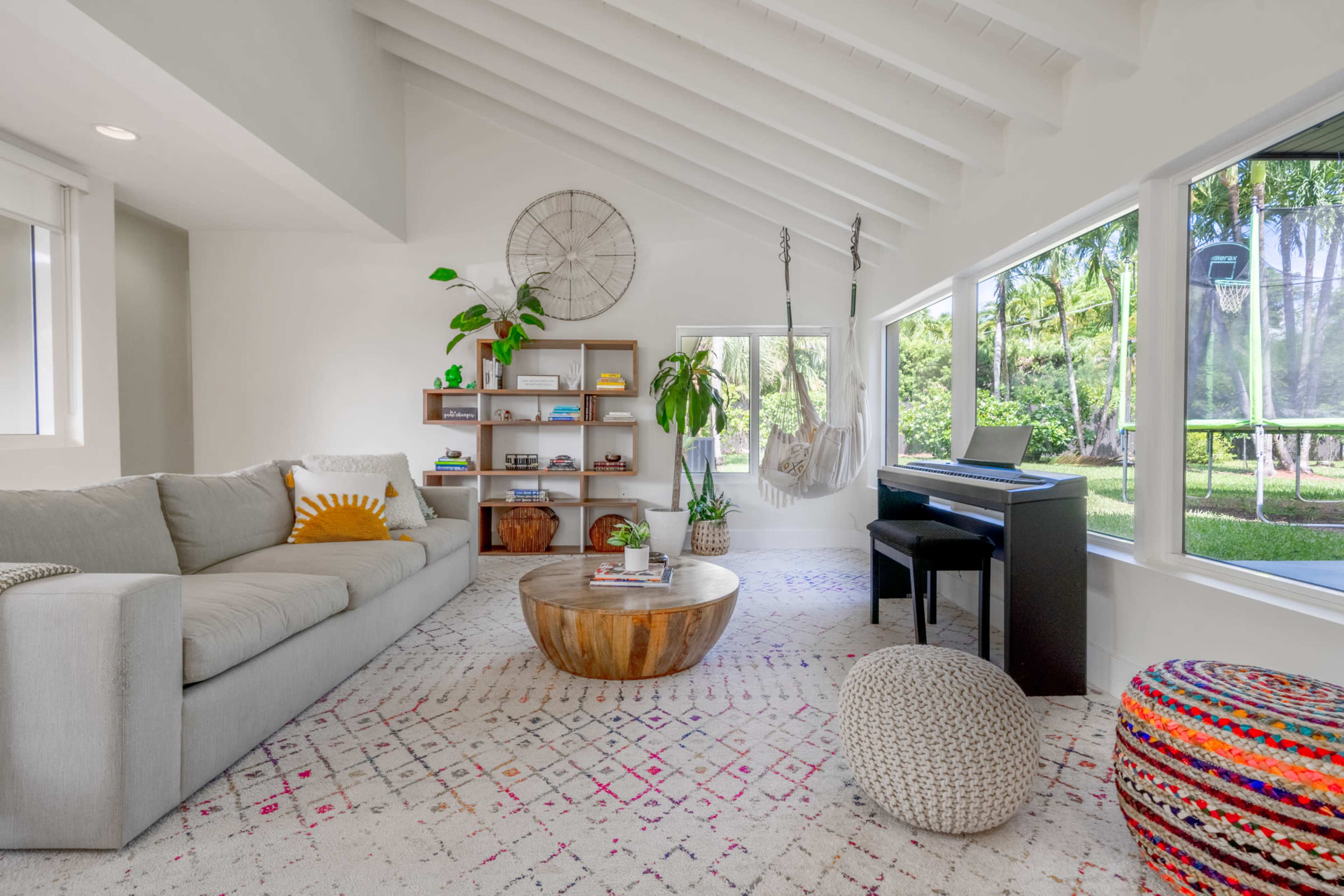 The living room features a light gray sofa, a wooden coffee table, a black piano, and large windows that reveal a view of a garden with a basketball hoop.