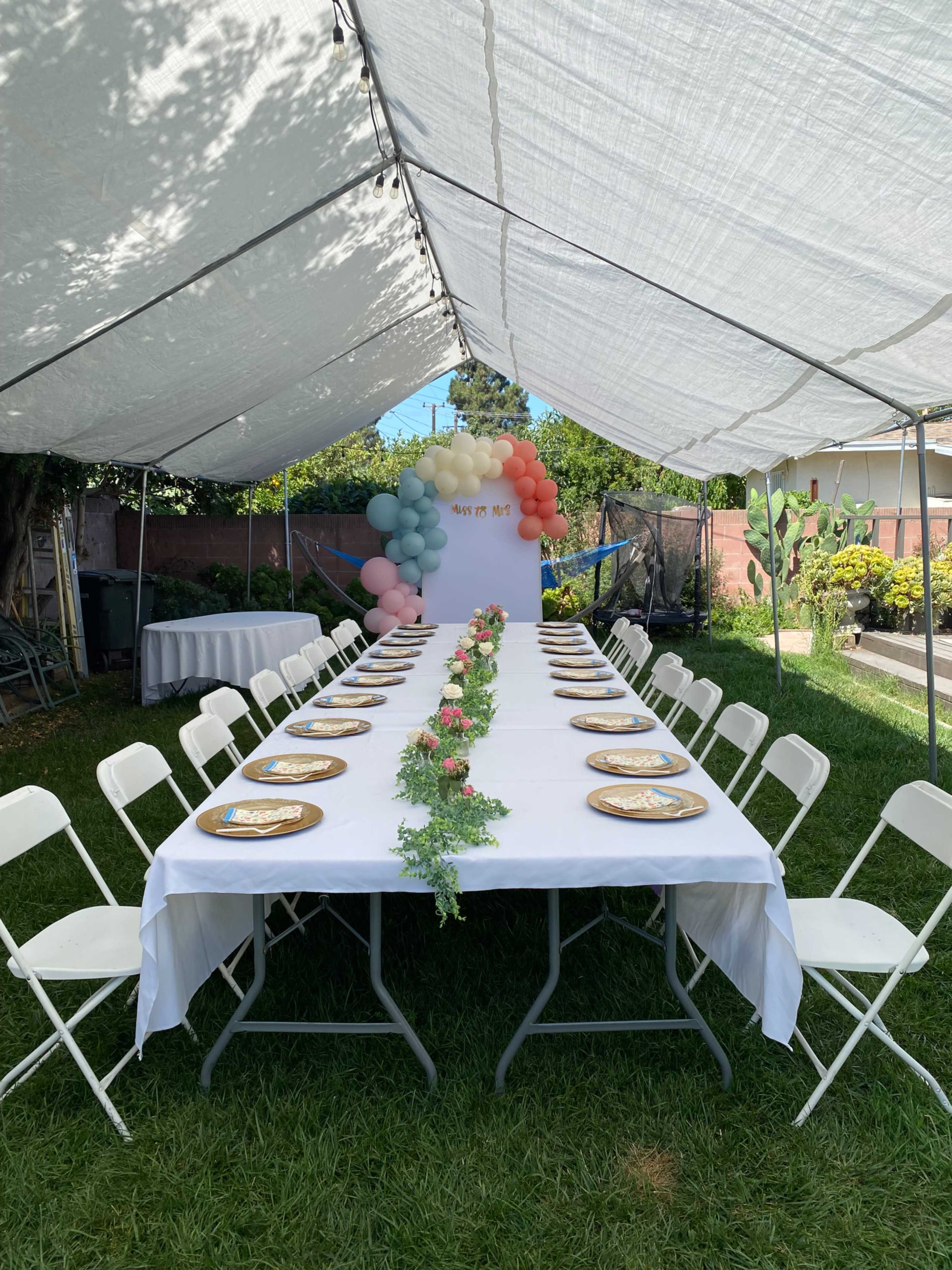 A long table is set under a tent, decorated with plates and a floral centerpiece, surrounded by chairs on a grassy area.