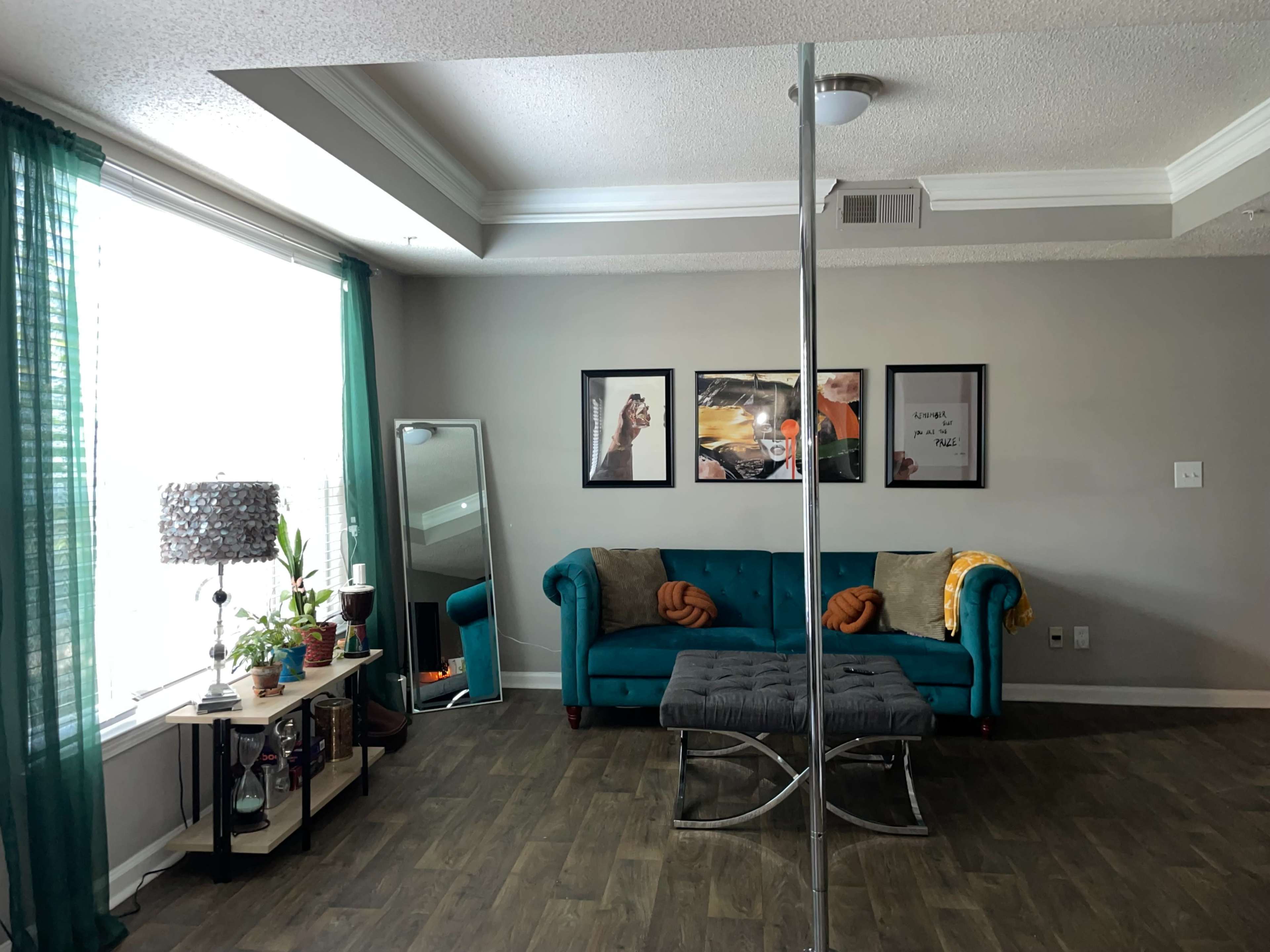 The image shows a living room with a turquoise sofa, a mirrored coffee table, and artworks on the wall, illuminated by natural light from a large window.