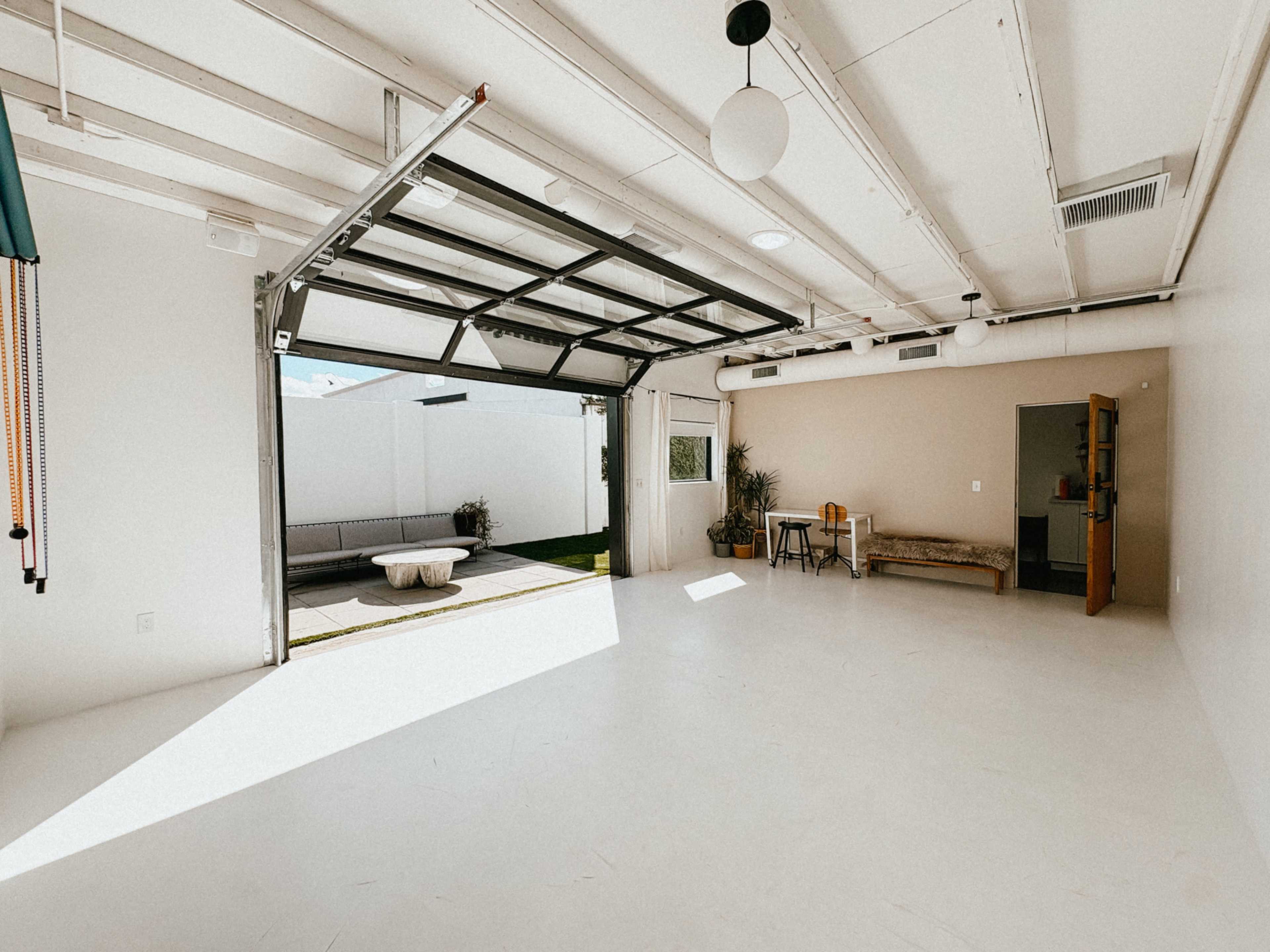 The image shows a spacious, empty room with a large garage door open, revealing a small outdoor area beyond.